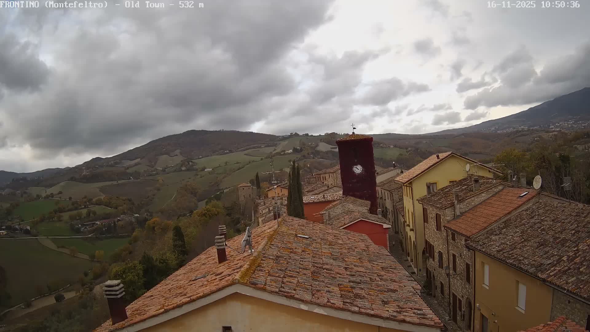 An aerial view overlooks an old European town with terracotta roofs and a prominent clock tower, set amidst rolling green hills under a cloudy, overcast sky.