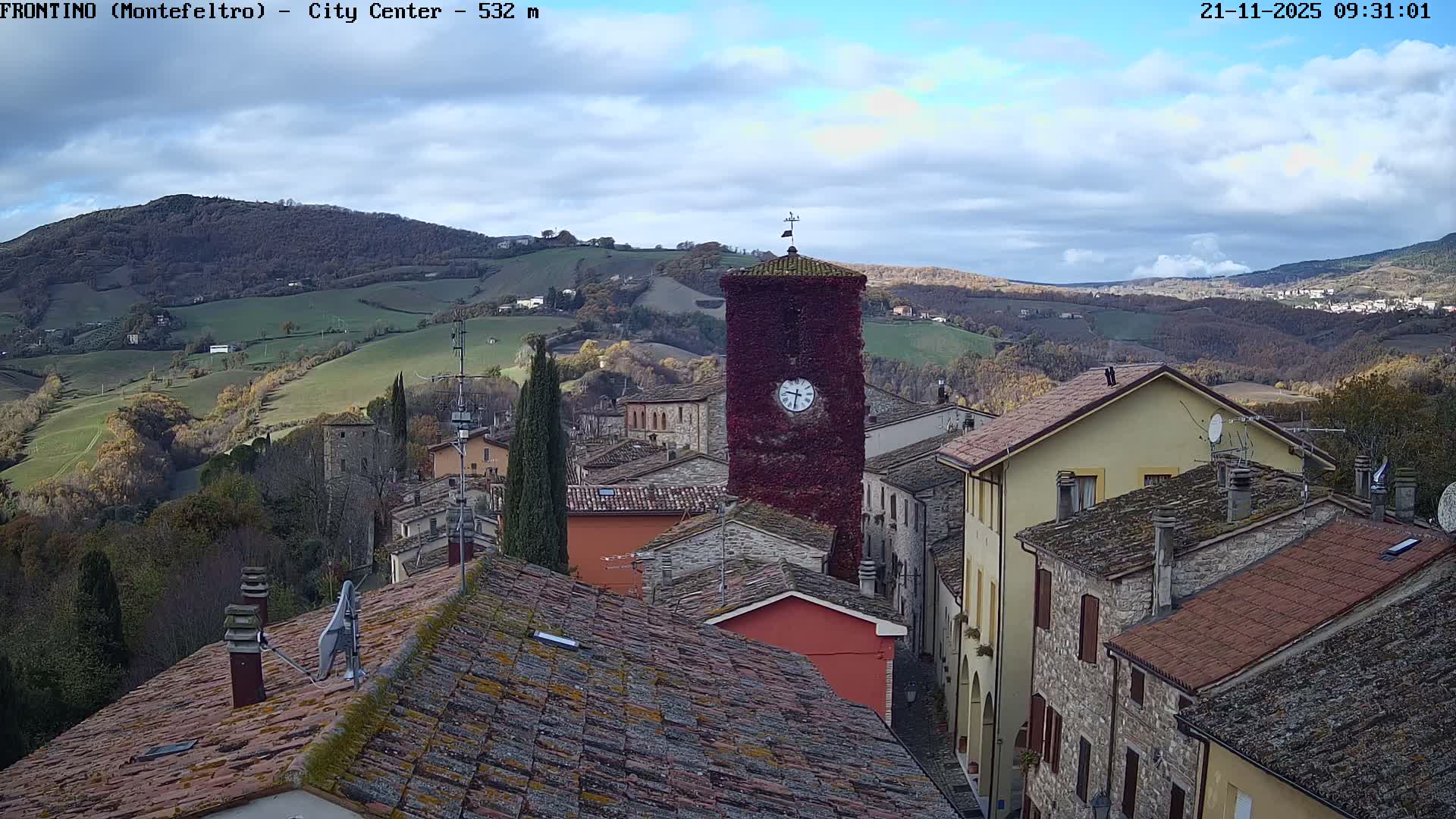 A scenic overlook captures a picturesque Italian village featuring a prominent ivy-covered clock tower and tiled rooftops, nestled amongst rolling green hills under a partly cloudy sky.