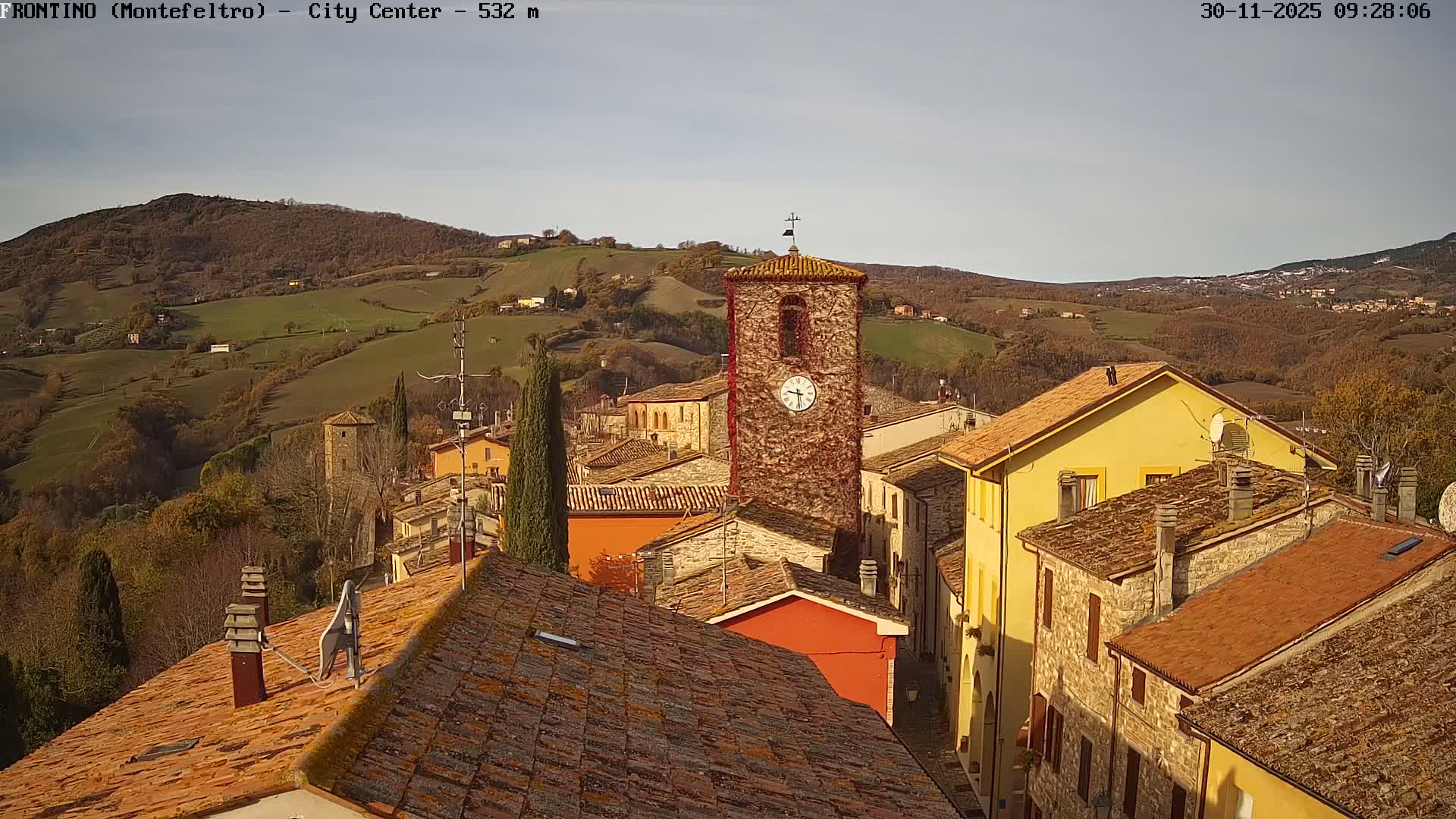 On a clear, sunny day, a picturesque village with a tall stone clock tower and houses featuring terracotta roofs is seen nestled within rolling hills displaying autumnal foliage.