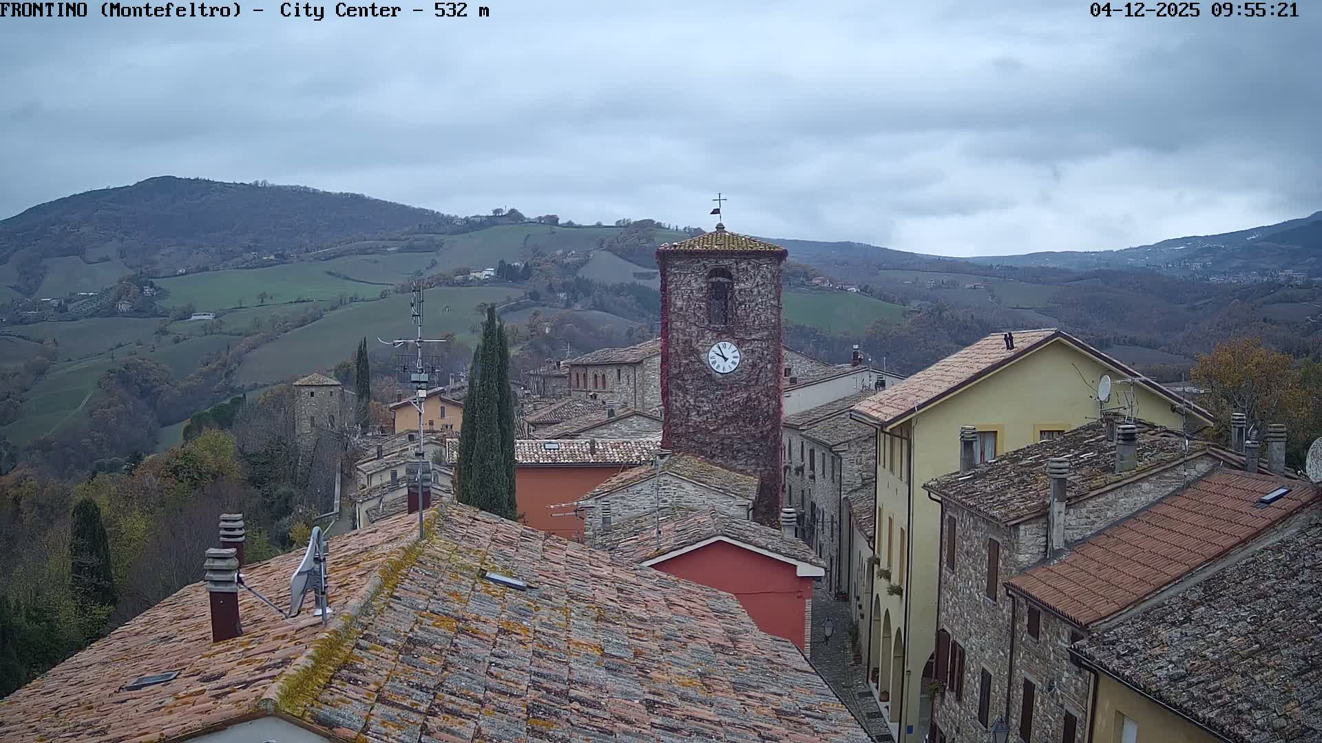 On an overcast day, an elevated view captures an old European hilltop town, featuring a prominent ivy-covered stone clock tower, terracotta-tiled rooftops, and cypress trees, with rolling, partially forested hills stretching into the distance.