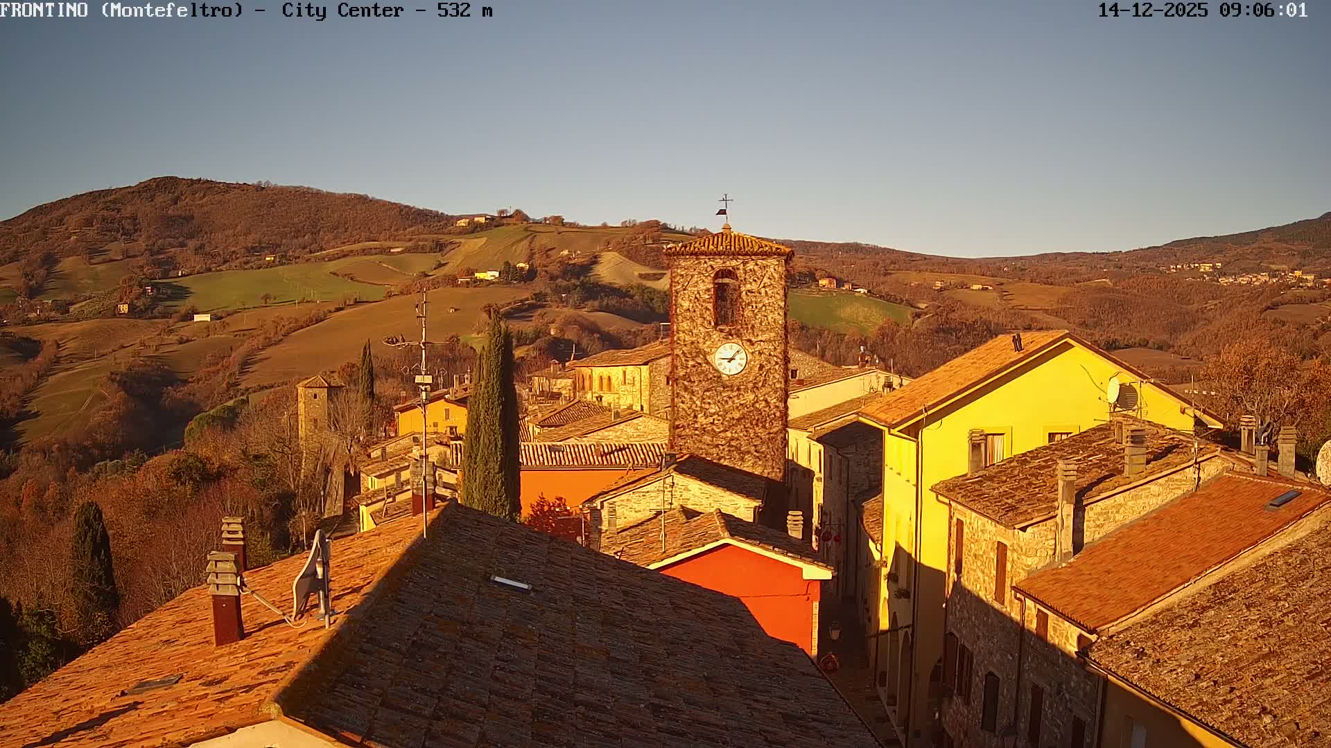 An elevated view of a sunlit Italian hill town features a prominent stone clock tower amidst traditional buildings with tiled roofs, set against a backdrop of rolling, autumnal hills under a clear blue sky.