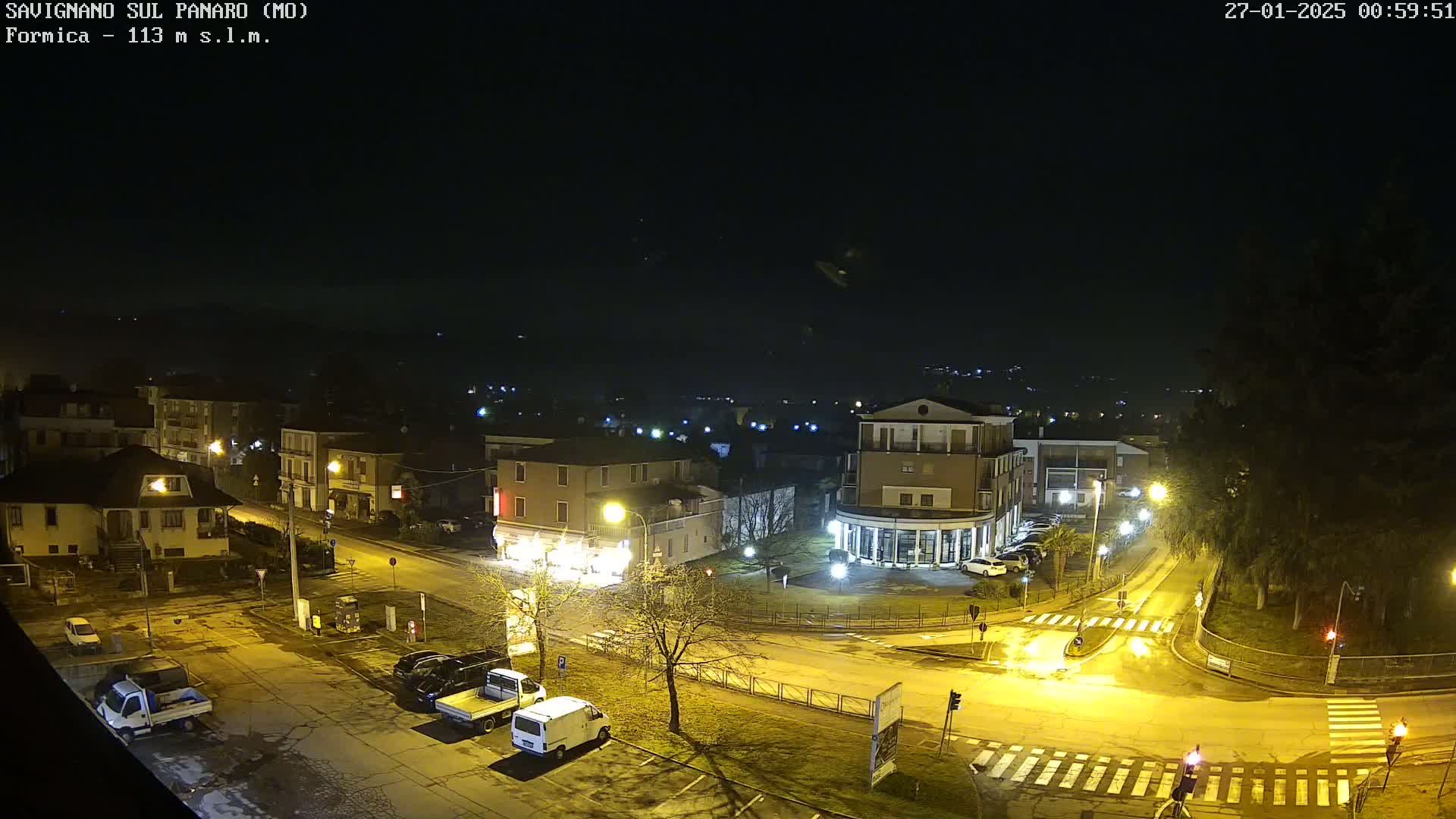 Here is a description of the image: A nighttime, high-angle view of a town square, showing several buildings, parked vehicles, and streetlights illuminating the scene under a clear, dark sky.