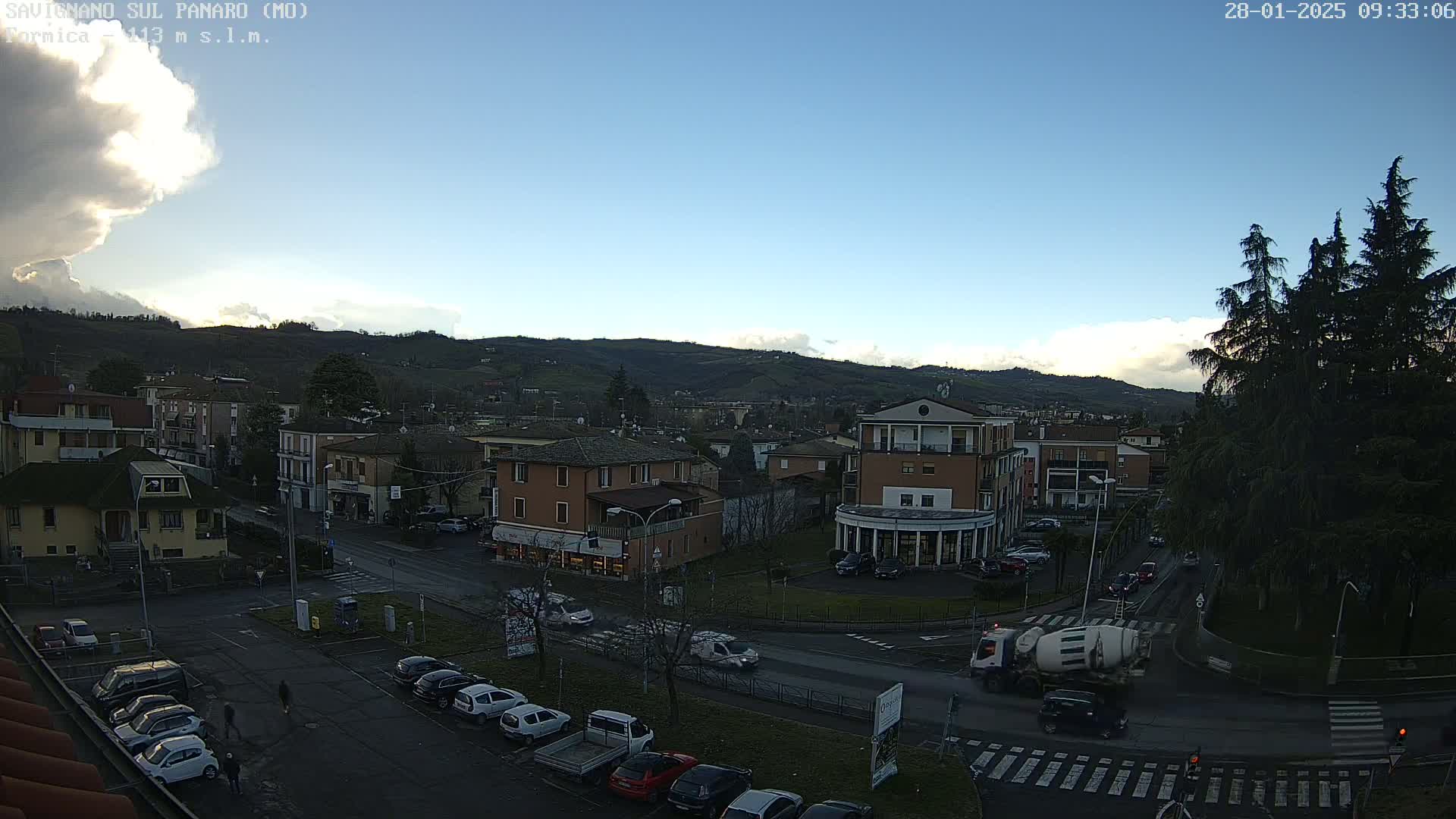 A partly cloudy daytime view of a town situated in a valley, with cars parked and driving on streets, and a cement mixer truck visible.