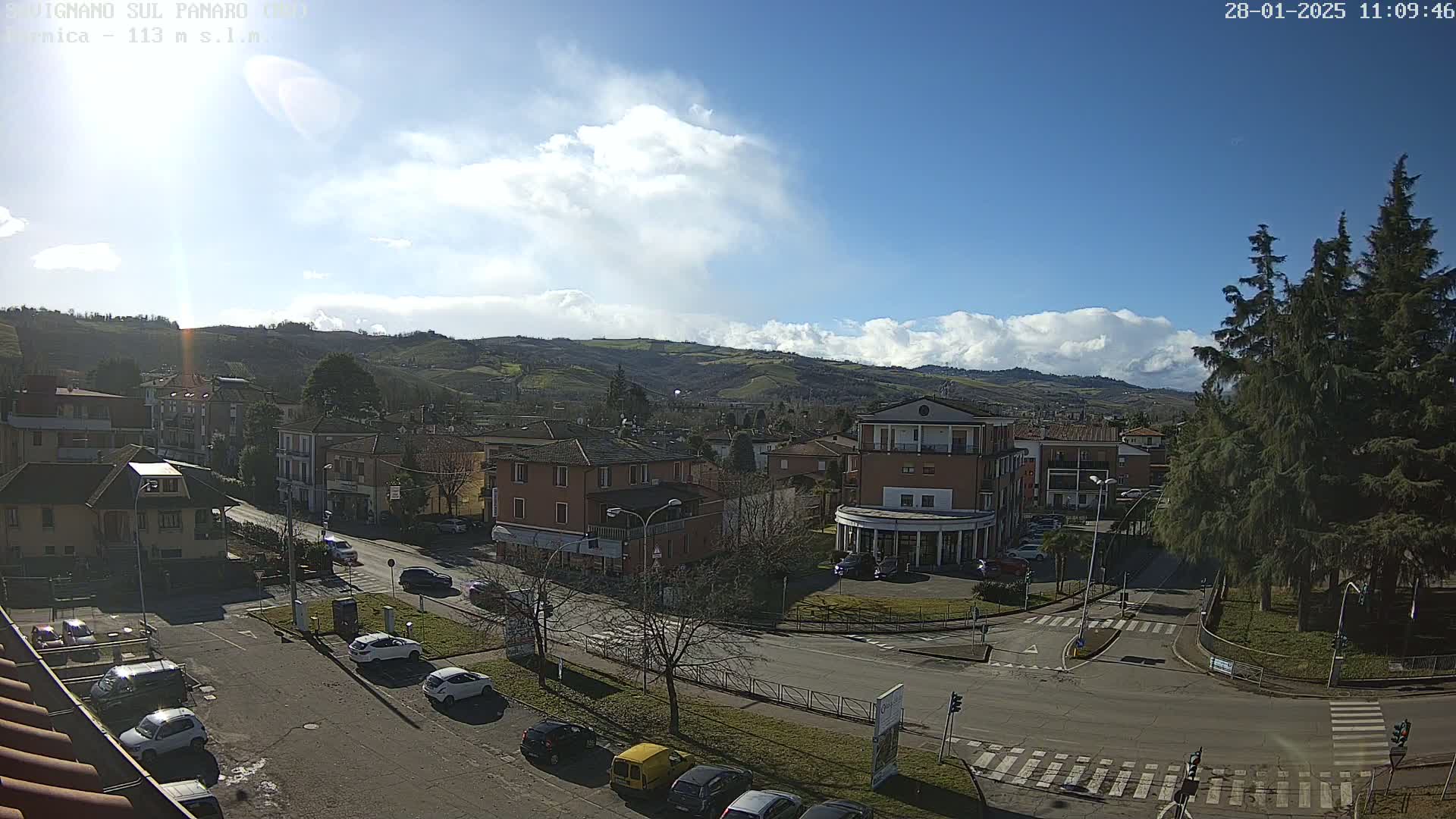 A sunny day with some clouds overhead reveals a view of a town nestled in a valley, with cars parked along streets and buildings visible.