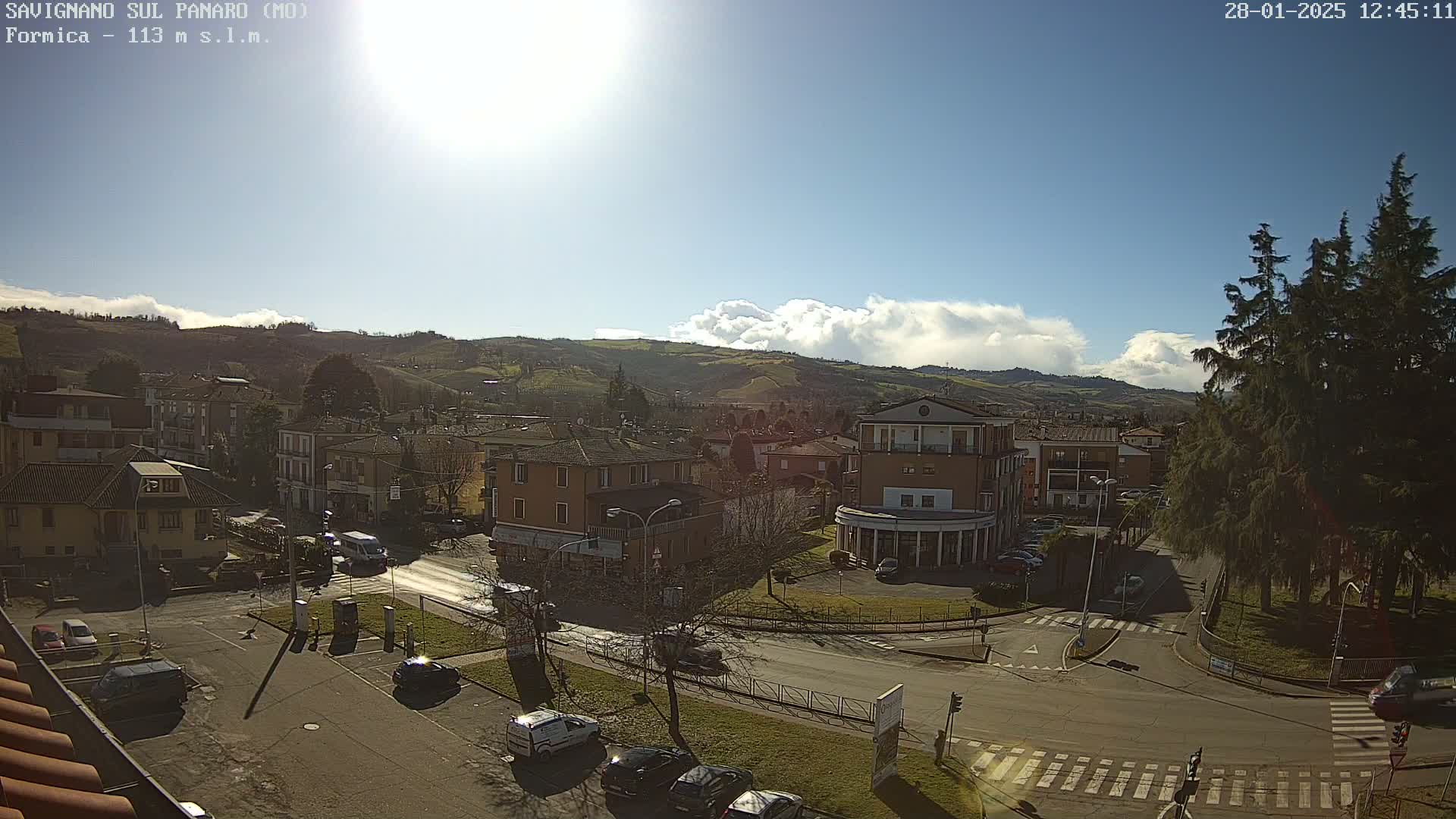 A sunny day reveals a view of a town nestled in a valley, with rolling hills and puffy clouds in the background.