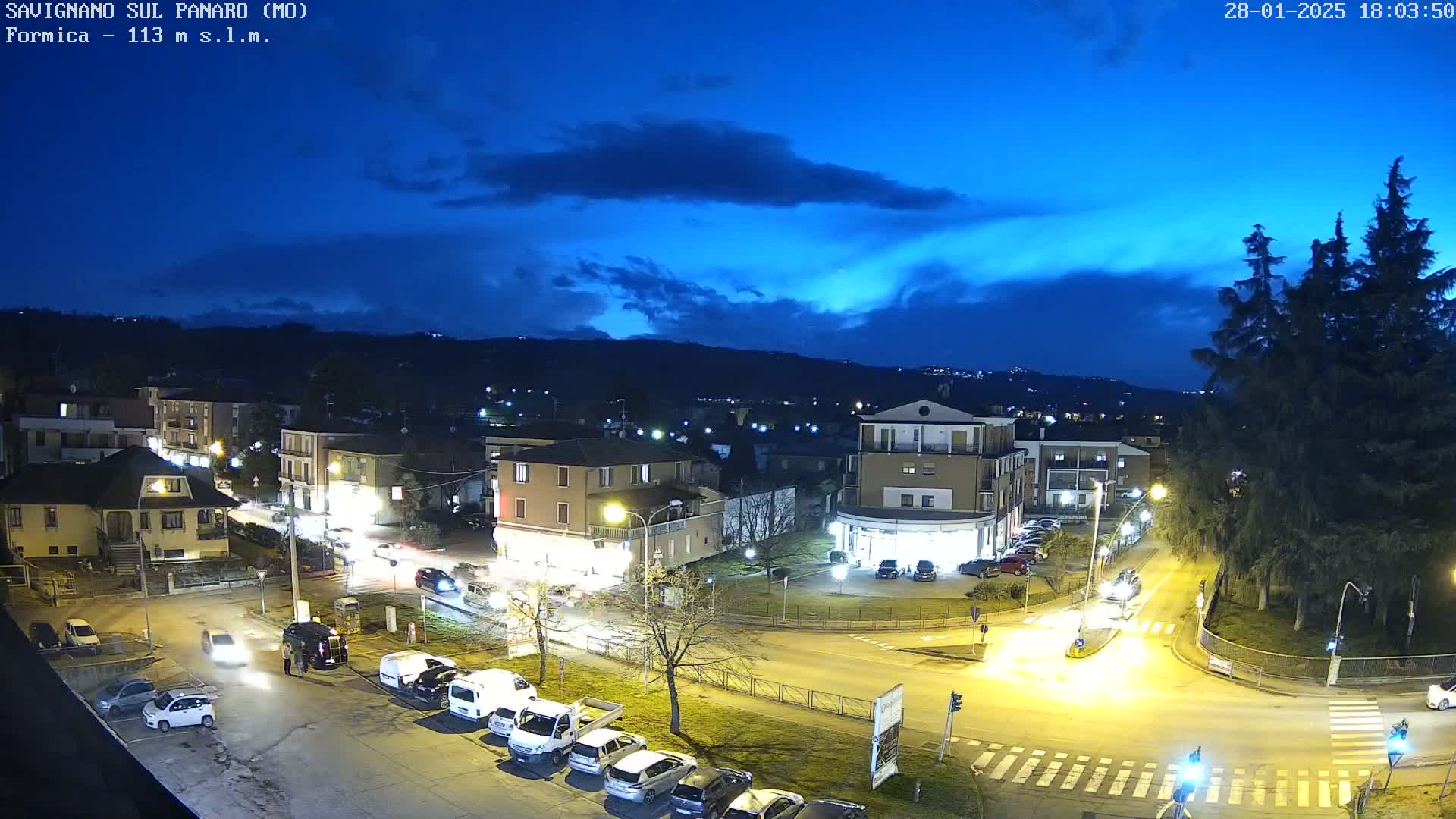 A nighttime aerial view of a town under a partly cloudy blue sky, showing streetlights illuminating cars parked and driving on roadways that intersect at a traffic circle.