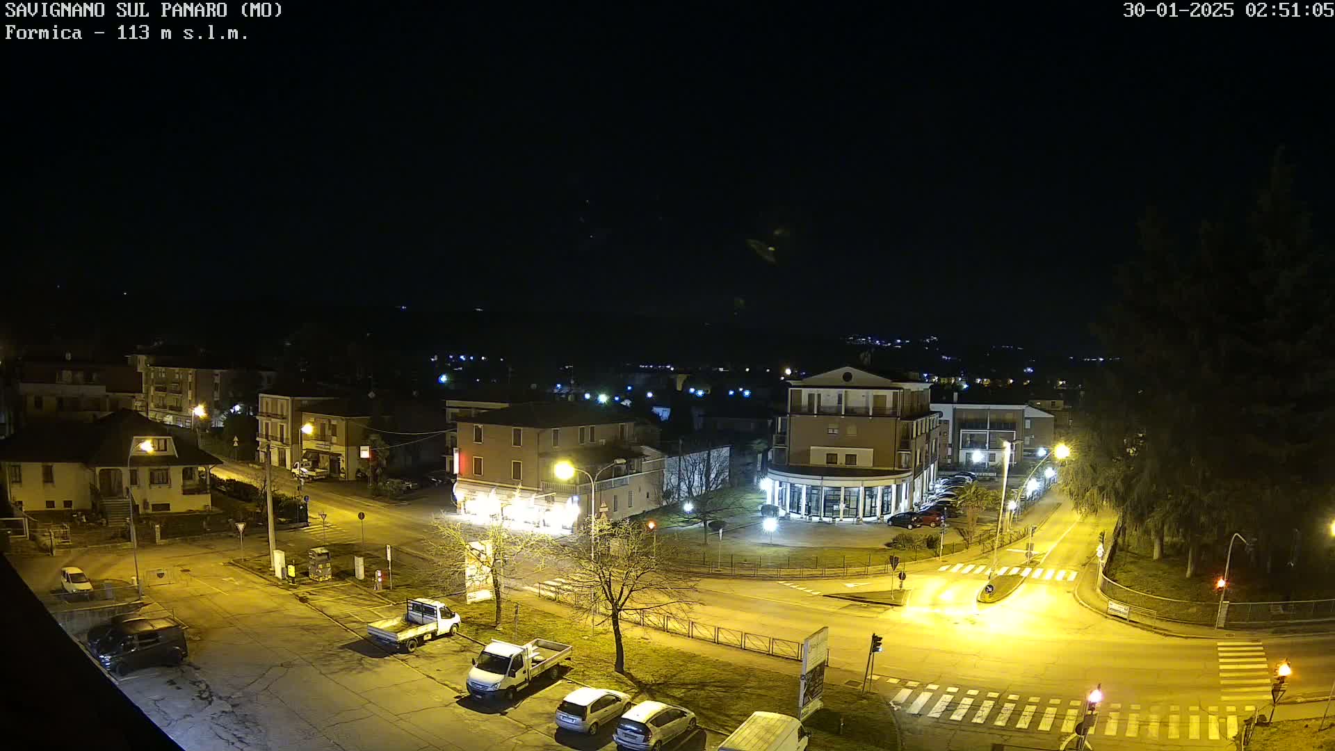 A nighttime aerial view of a town square with several cars parked and driving through the intersection, under a clear, dark sky.