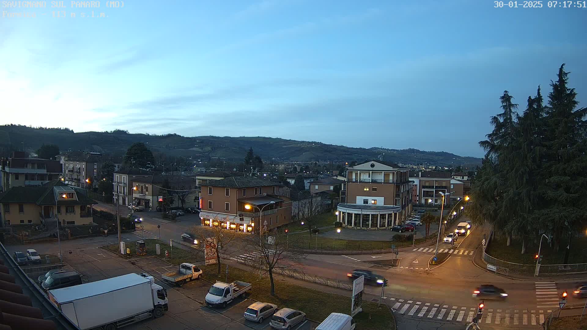 A twilight view of a town square with cars and trucks in the parking lots and on the roads, nestled beside rolling hills under a clear, dusky blue sky.