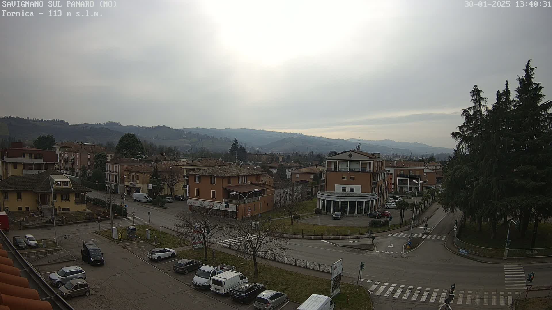 An overcast day reveals a view of a town square with parked cars, buildings, and a few trees, set against a backdrop of rolling hills.