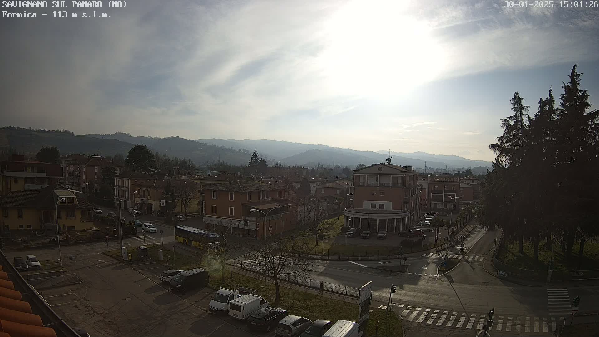 A partly sunny day reveals a town nestled in a valley, surrounded by hills and featuring a main intersection with cars and a bus.