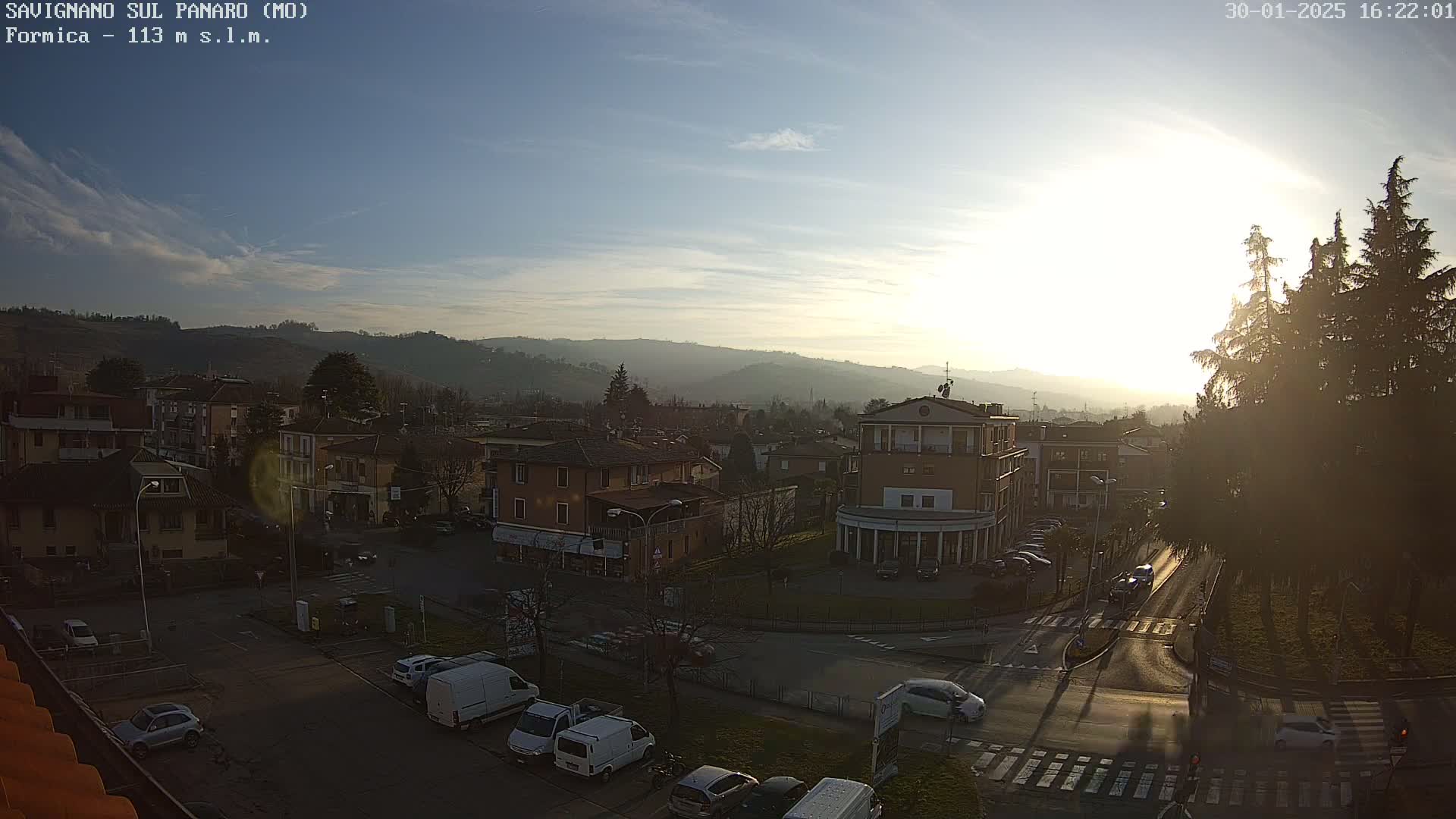 A sunny, partly cloudy day overlooks a town with low-lying buildings and hills in the background, cars parked along roadways and a few cars driving.