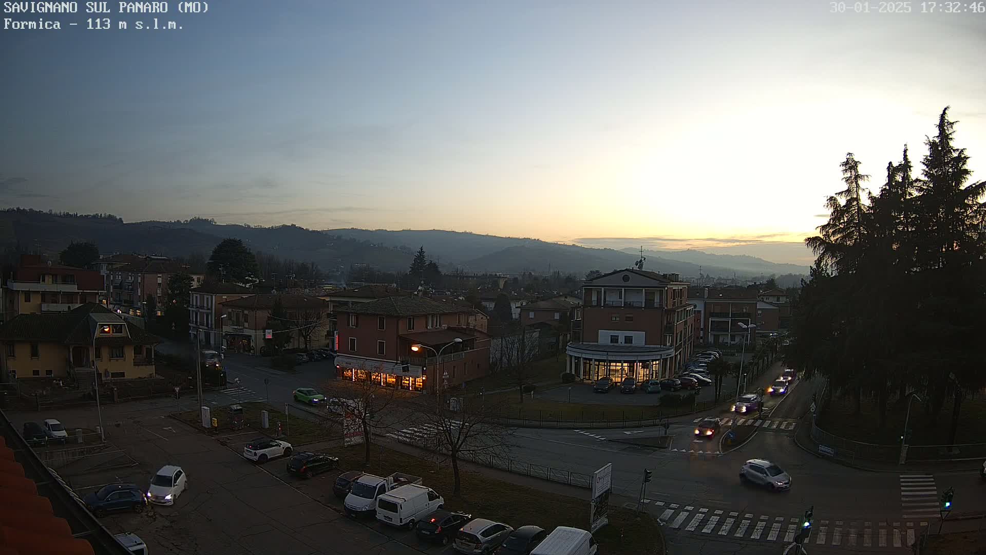 An aerial view of a town at dusk shows cars driving on roads that intersect near a small square with shops, set against a backdrop of hills under a clear, twilight sky.