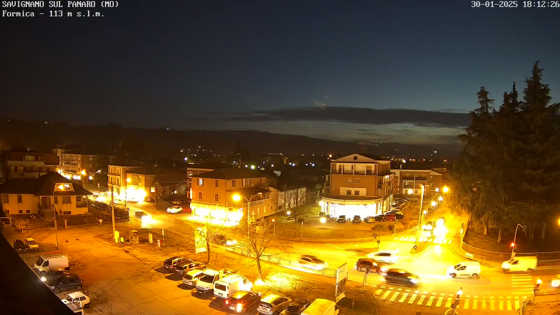An aerial nighttime view of a town square with cars driving around a roundabout, illuminated by streetlights under a mostly clear sky.