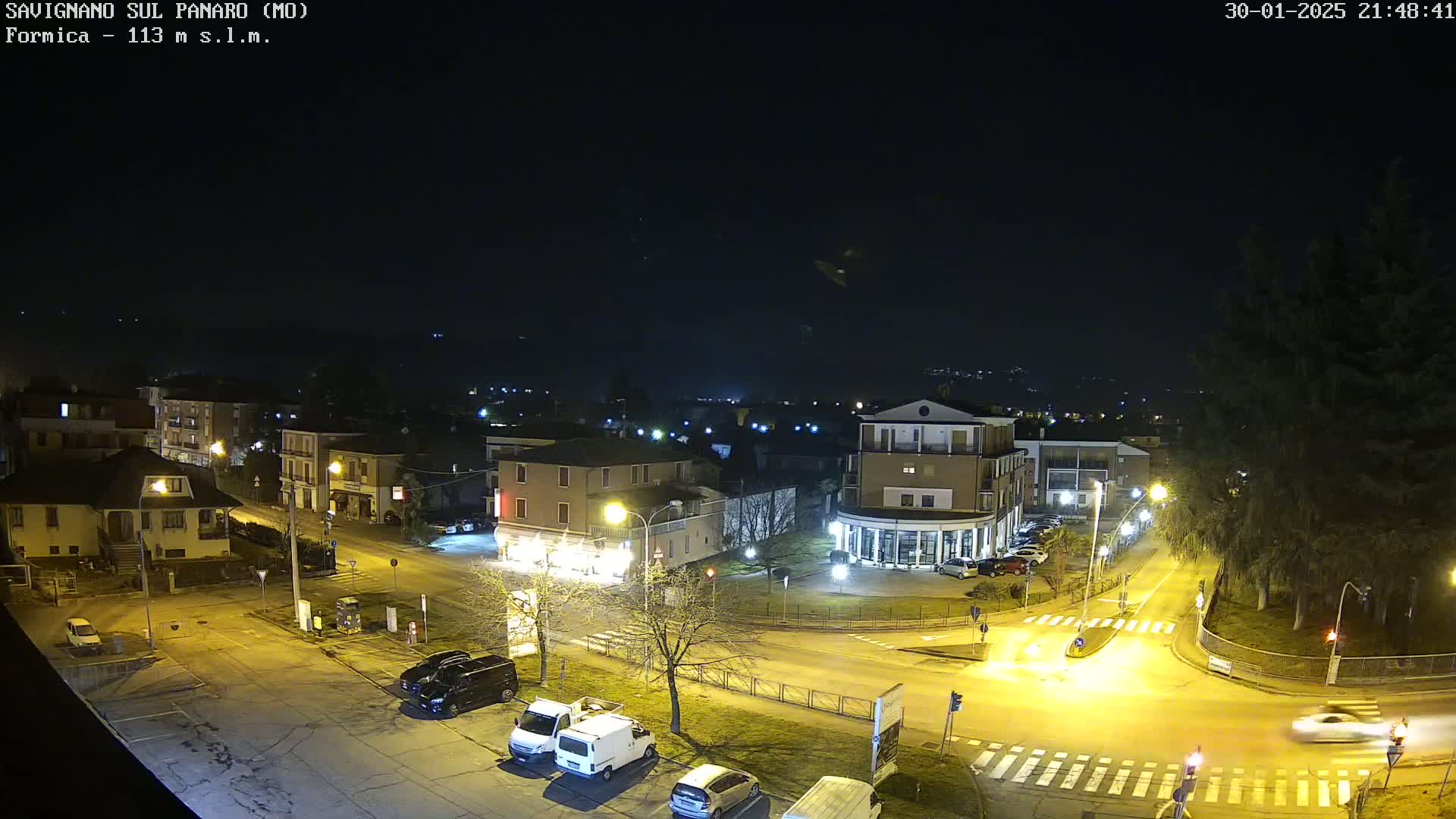 A nighttime, high-angle view of a town square with several cars parked and driving, illuminated by streetlights.