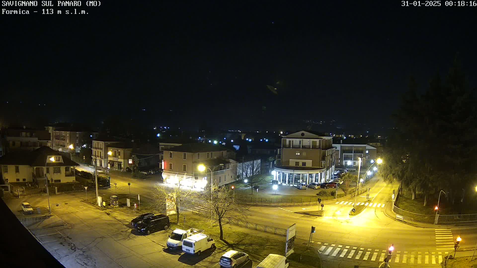 A nighttime aerial view of a town square with several buildings and vehicles, illuminated by streetlights under a clear, dark sky.
