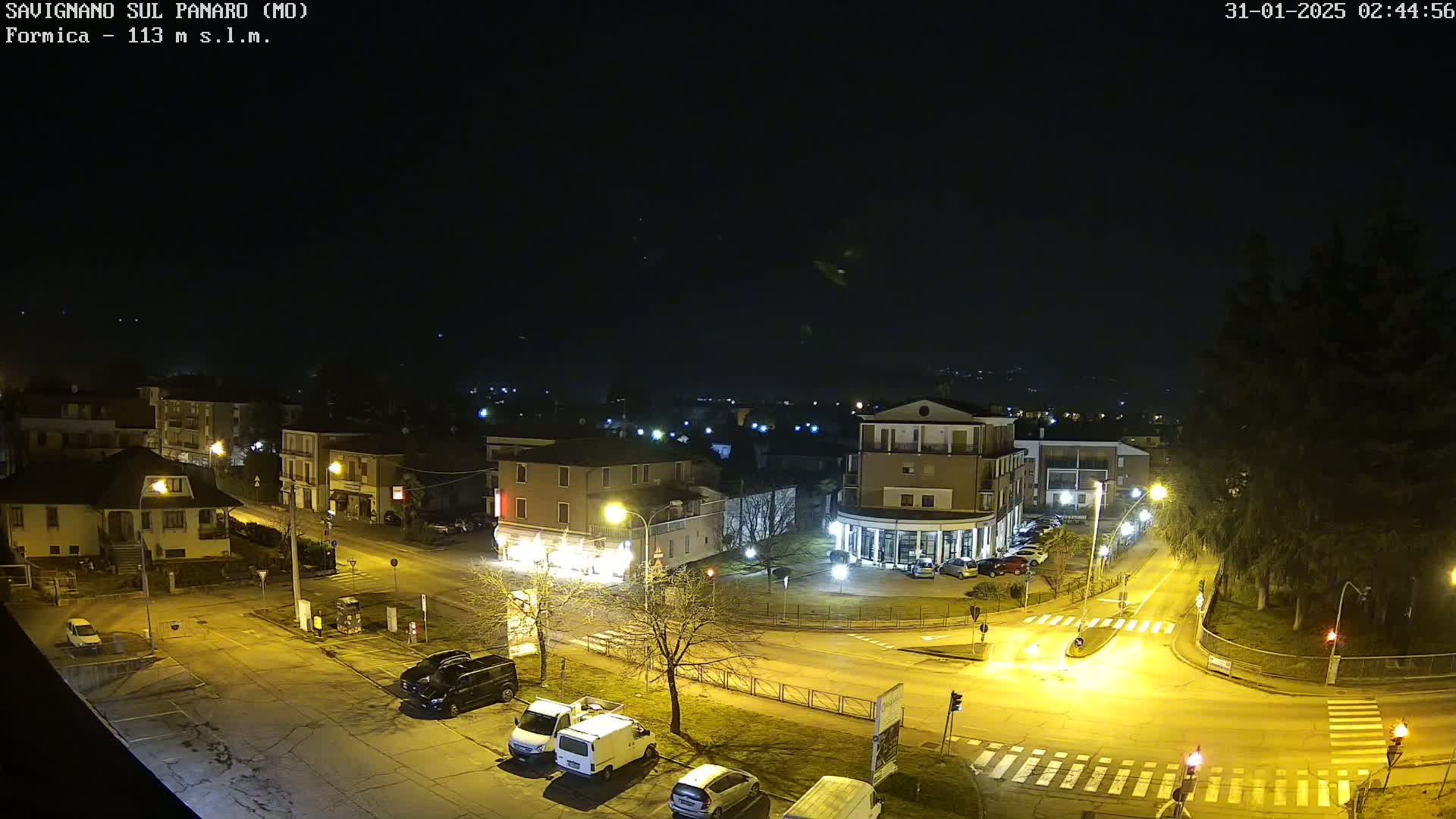 A nighttime, high-angle view of a town square with several buildings, parked cars, and illuminated streets.