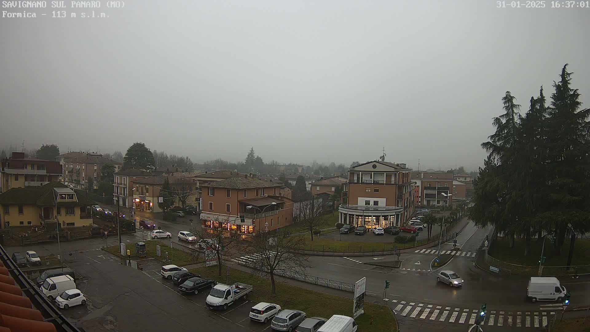 A misty overcast day reveals a town square with cars parked along the streets and several buildings, including a few two-story structures.