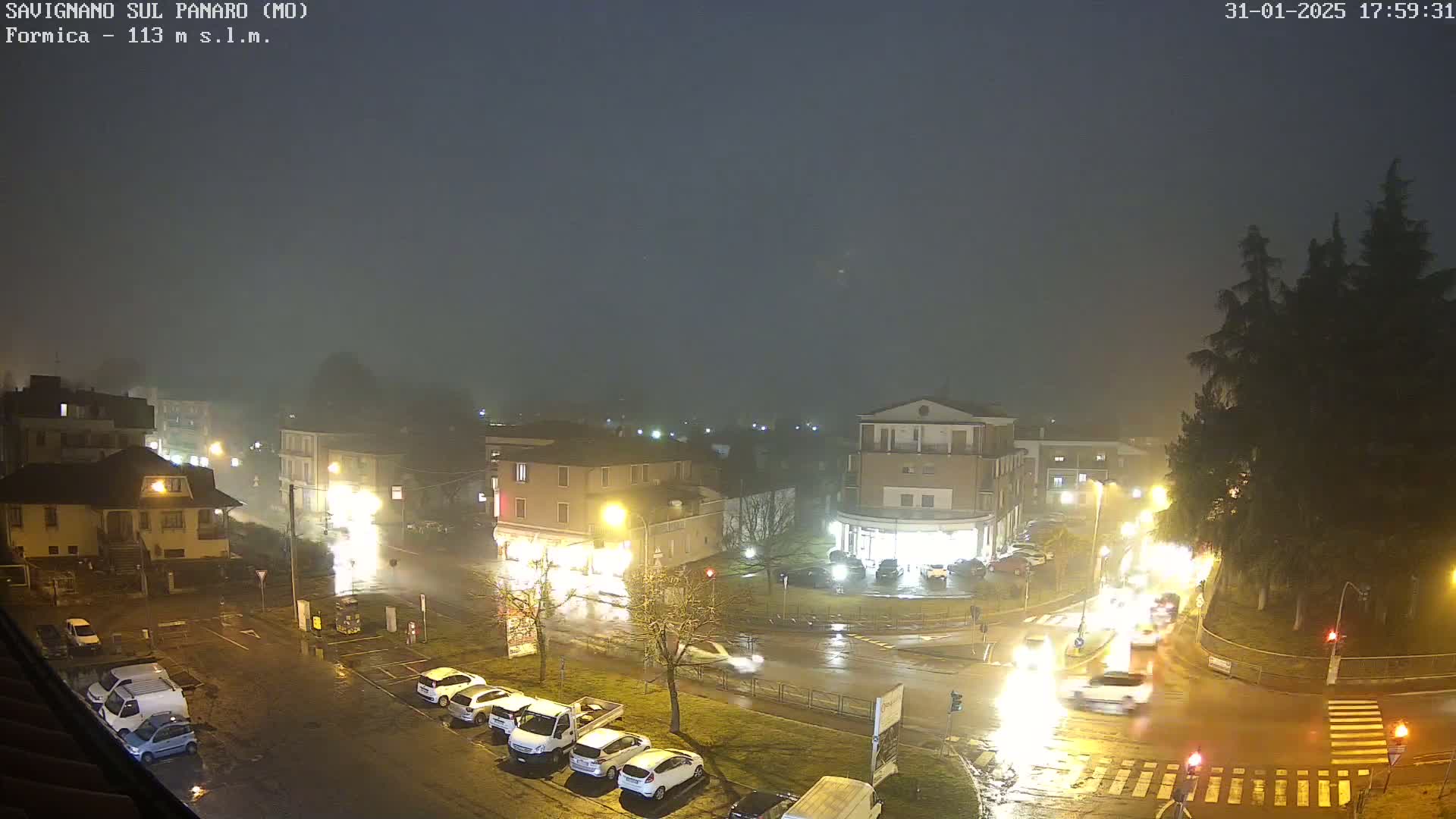 A nighttime, foggy view of a wet, rain-slicked town square with several cars and buildings illuminated by streetlights.