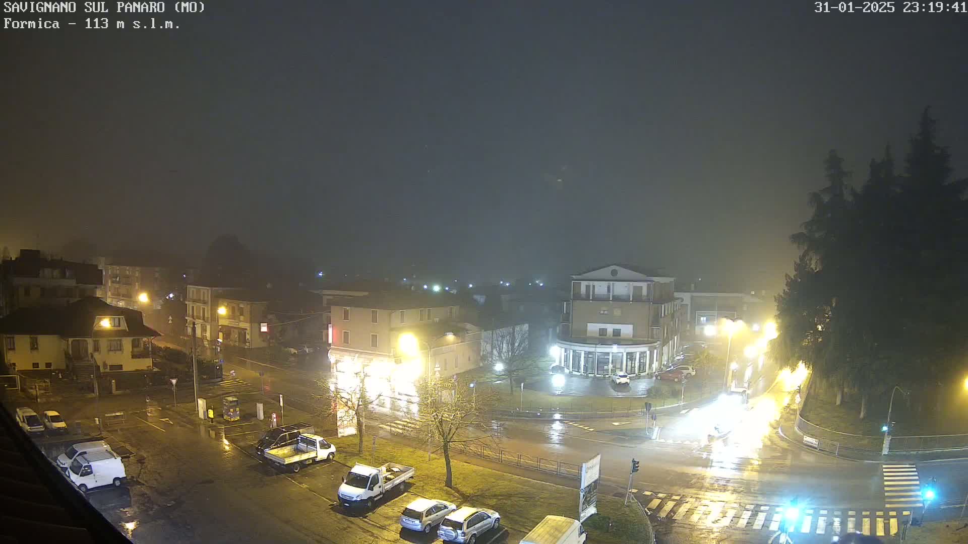 A foggy, rainy night scene shows a town square with parked cars and streetlights illuminating wet roads.