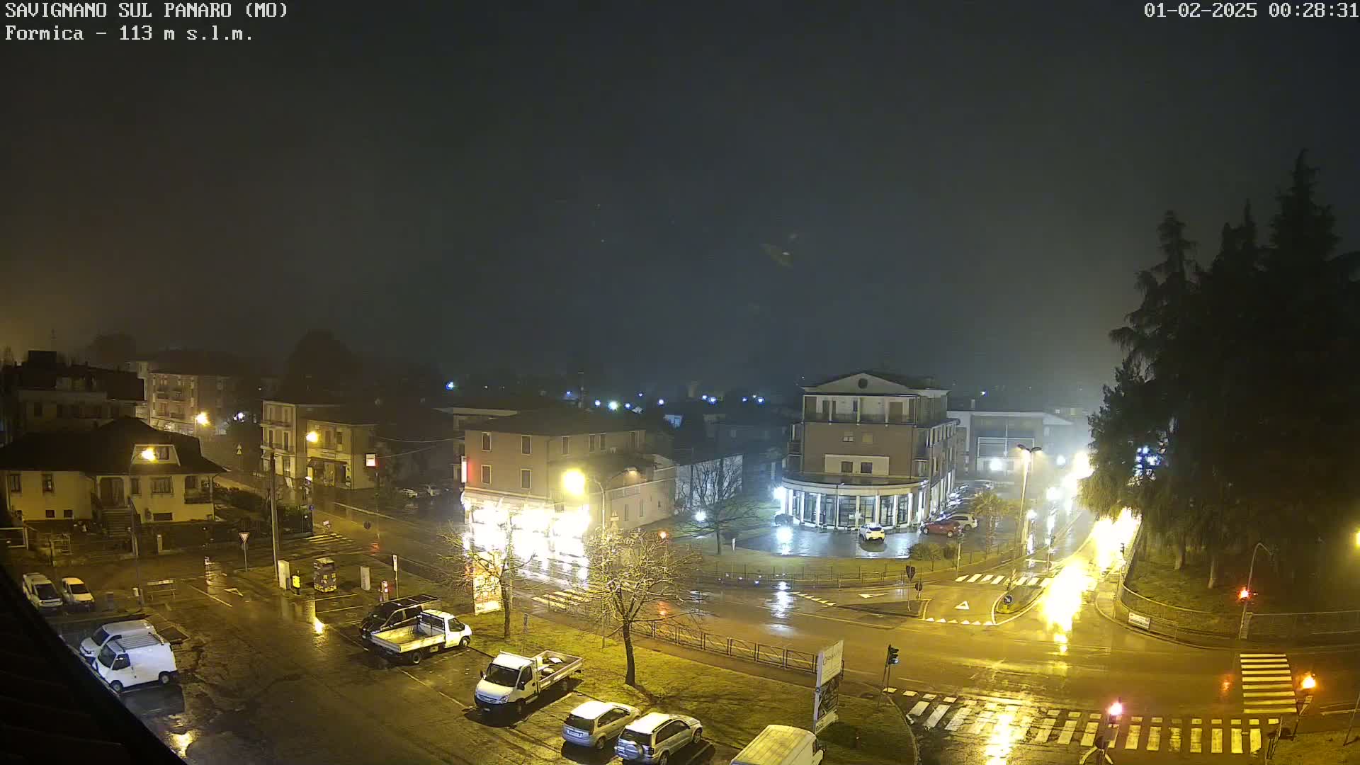 A wet, nighttime view of a town square with several buildings, parked cars, and streetlights reflecting on the rain-slicked streets.