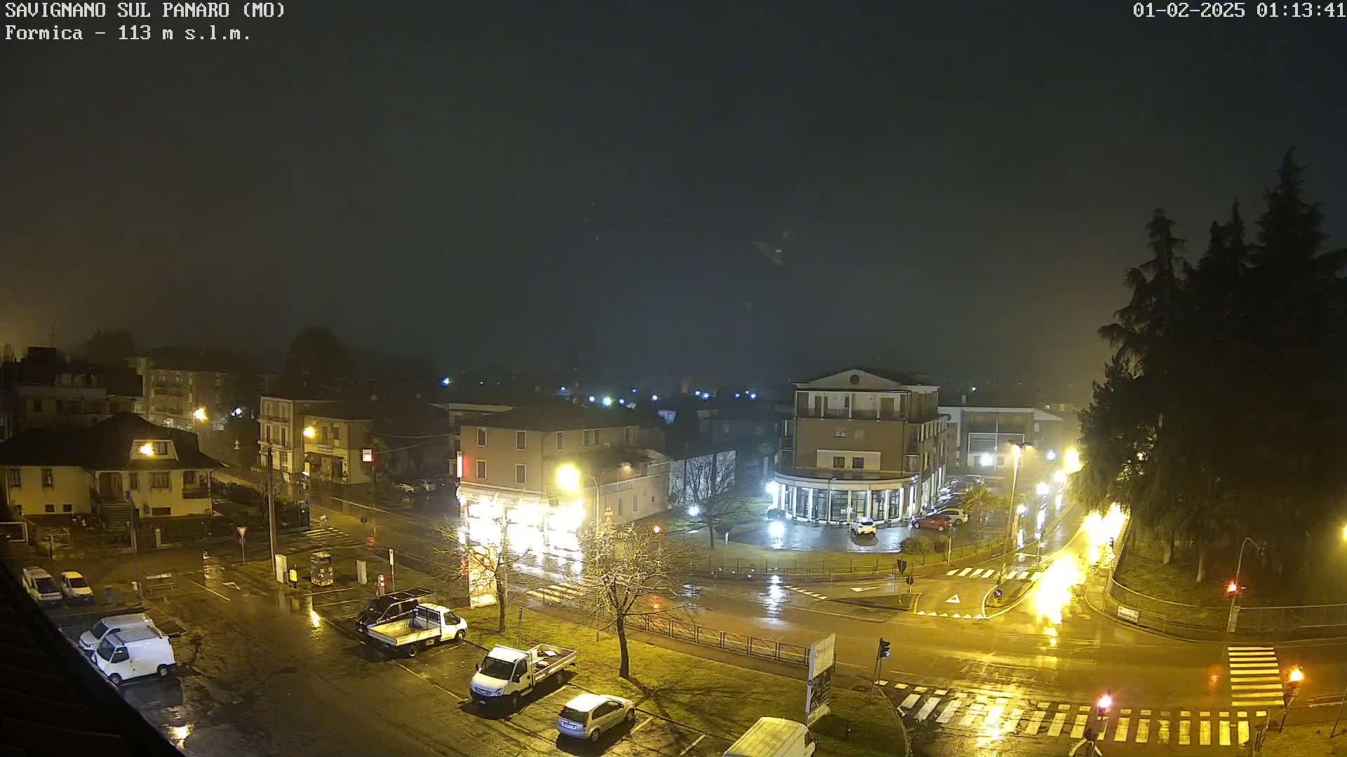 A nighttime, rainy view of a town square with parked cars and buildings illuminated by streetlights.