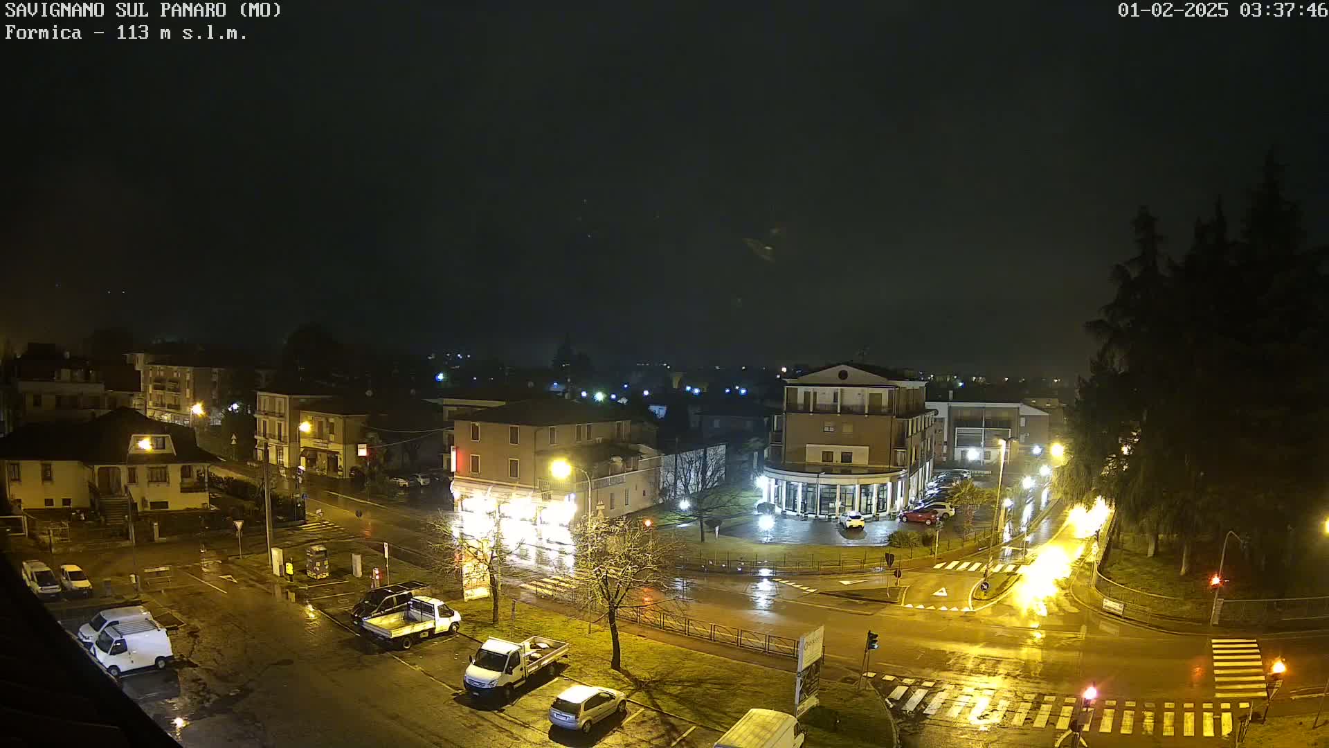 A nighttime, rainy view of a town square with parked cars and buildings illuminated by streetlights.
