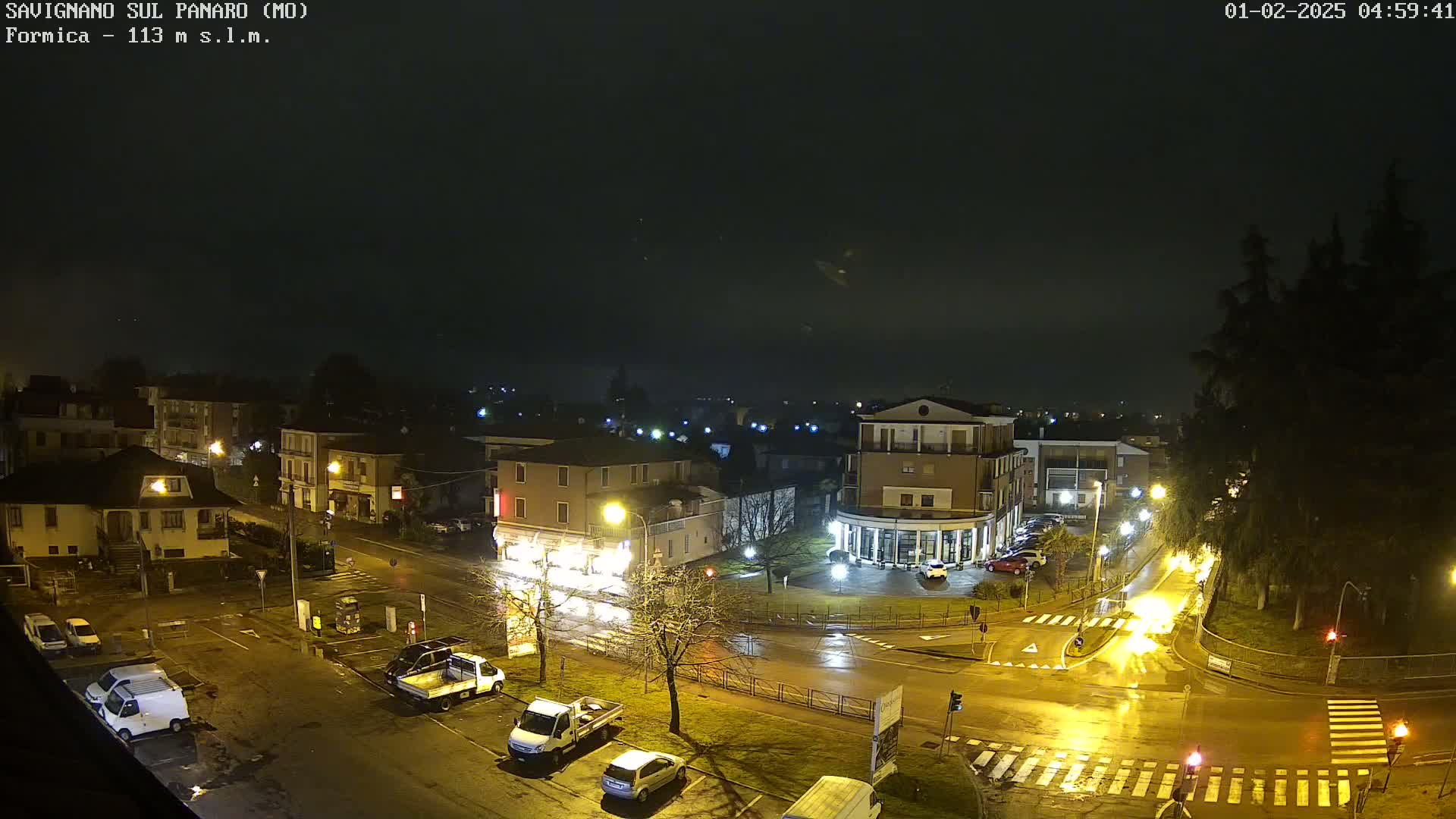 A wet, nighttime view of a town square with several cars parked and driving on wet roads under dim streetlights.
