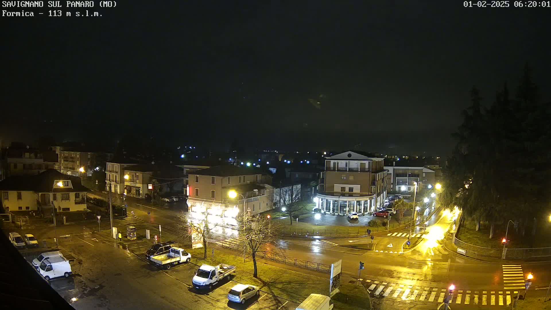 A wet, nighttime view of a town square with several buildings, parked cars, and streetlights reflecting on the rain-slicked roads.
