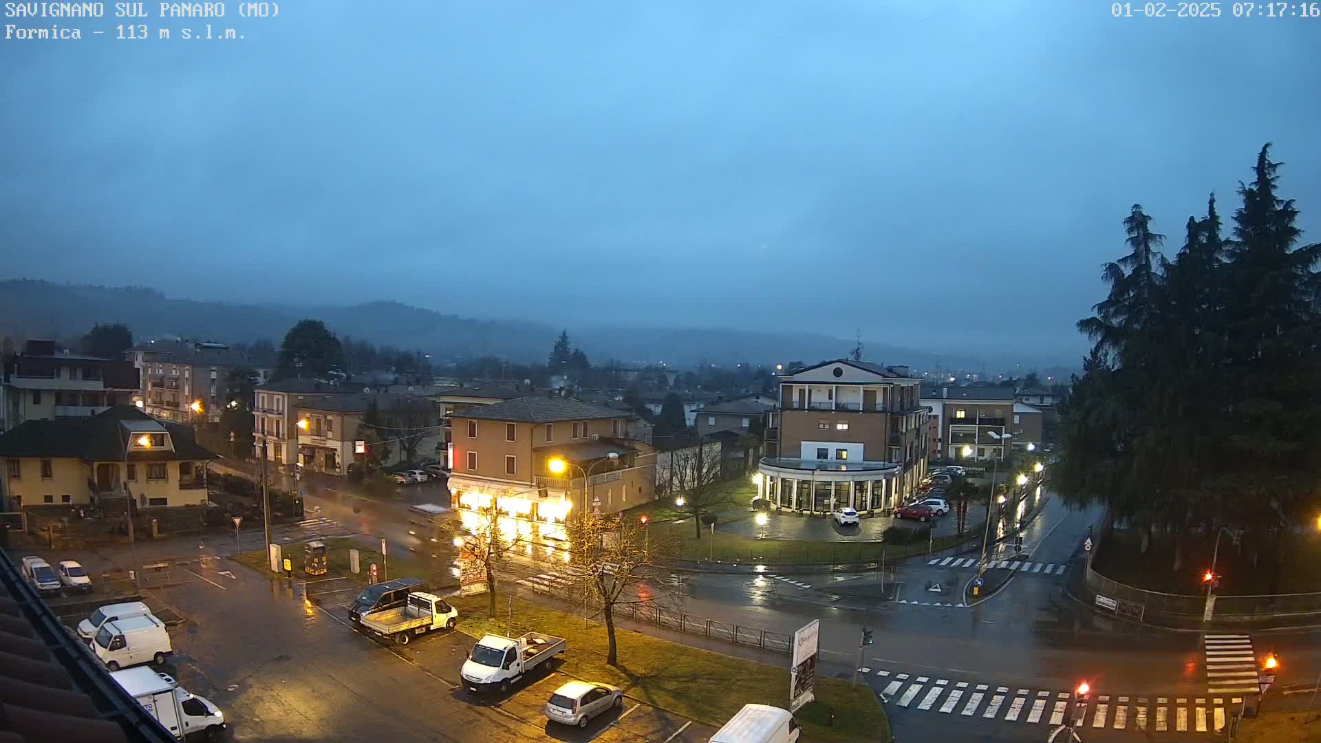 A rain-soaked town square, illuminated by streetlights at dusk, is surrounded by buildings and trees under a dark, overcast sky.