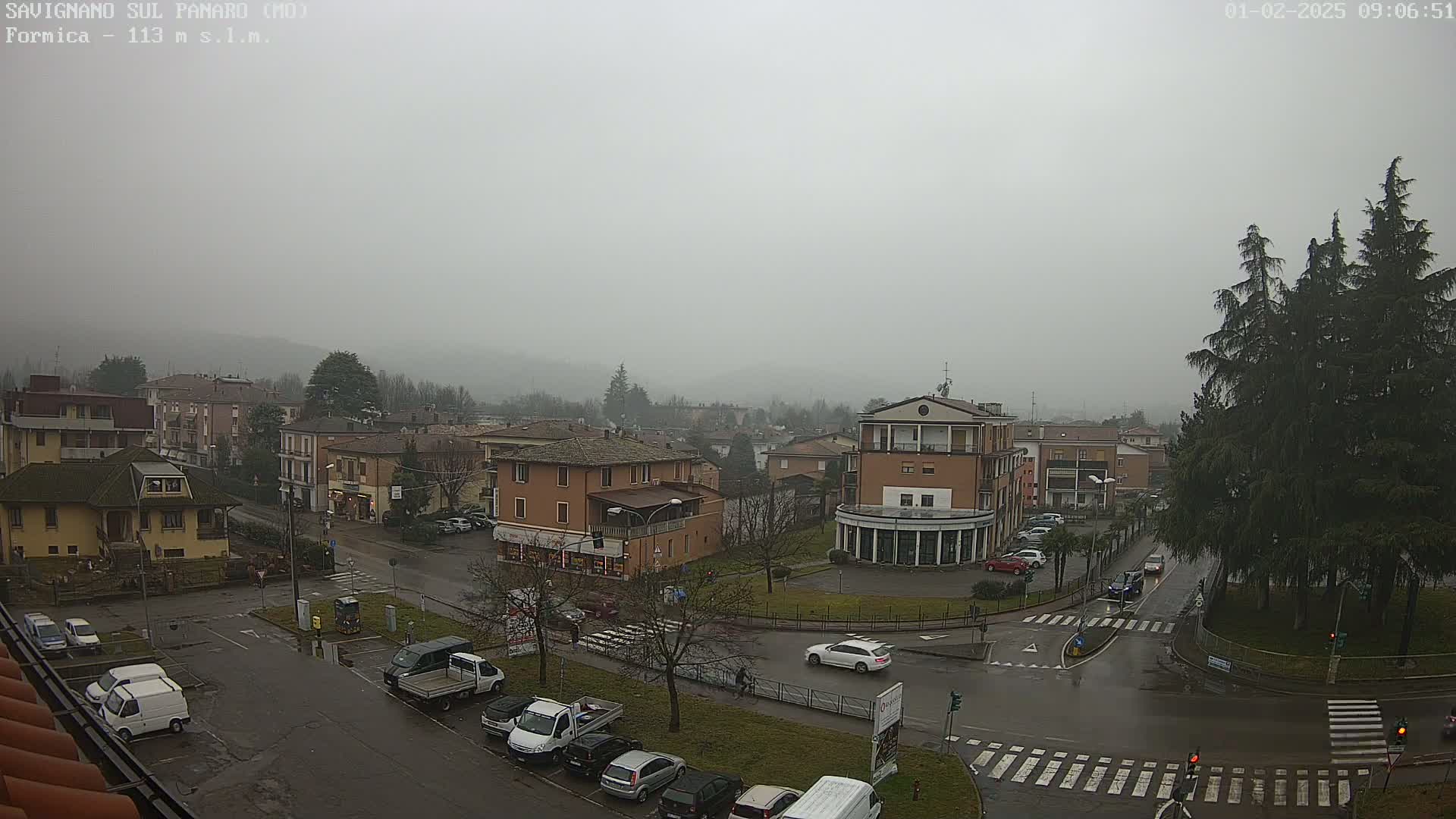 A foggy, overcast town square with several buildings, cars parked in lots, and a few cars driving on wet roads.