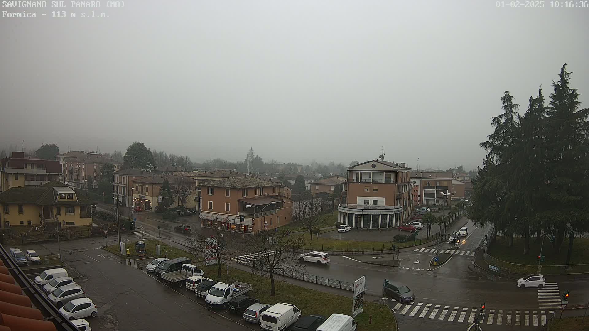 A foggy, rainy day reveals a street intersection in a town with several buildings, cars parked along the sides of the roads, and tall evergreen trees.