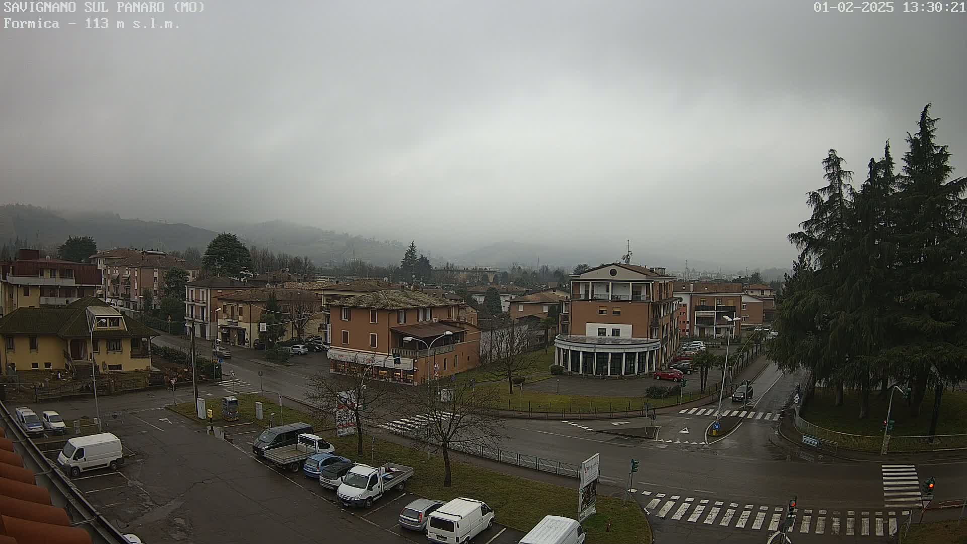 A foggy, overcast view of a town square with several buildings, parked cars, and a few sparse trees, showing wet roads.