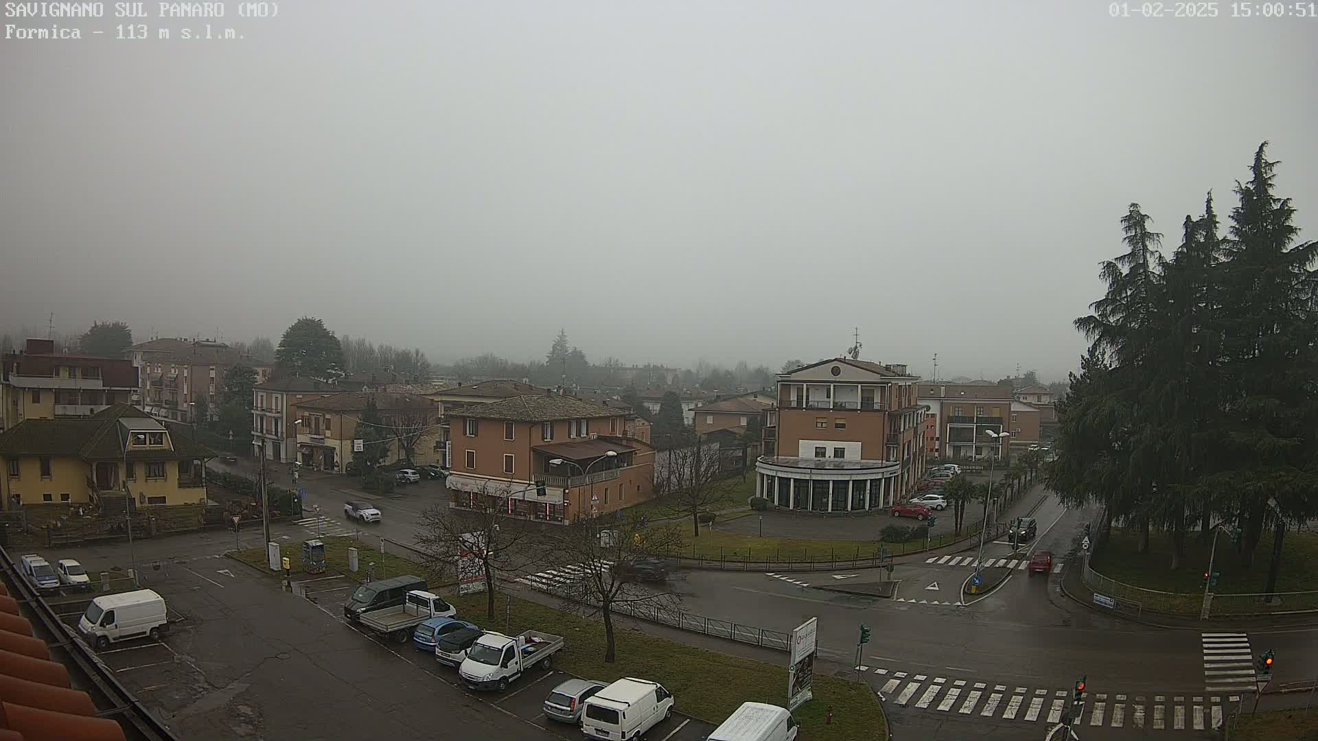 A misty view of a town square with several buildings, parked cars, and a few cars in transit, under a gray sky.