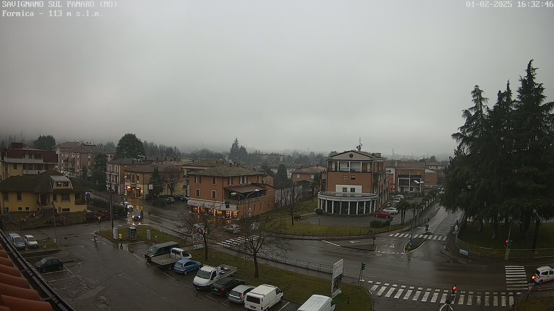 A wet, overcast town square with several buildings and cars parked along the roads.
