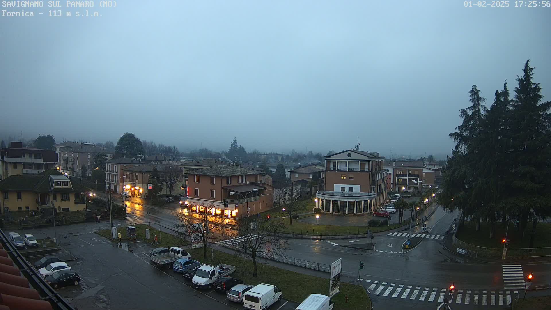 A misty, rain-slicked town square with cars parked in lots and a few vehicles on the roads, surrounded by low-rise buildings under a dusky sky.
