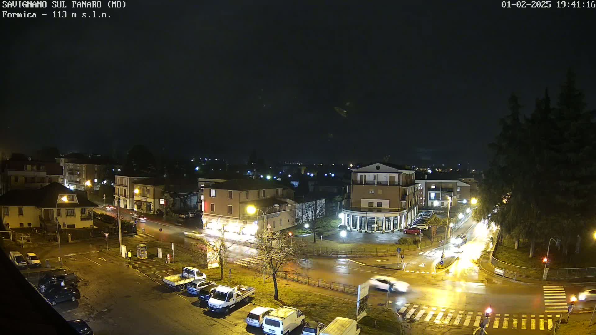 A nighttime, wet view of a town square with cars and buildings illuminated by streetlights under a dark, cloudy sky.
