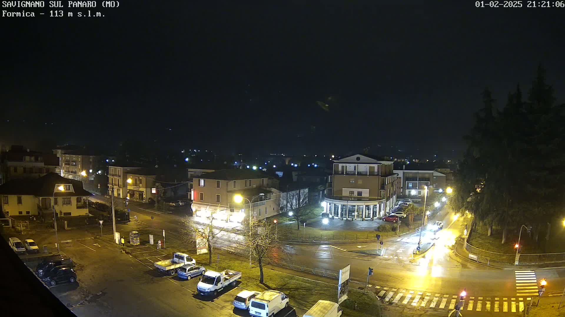 A wet, nighttime view of a town square with cars parked and driving around a roundabout, illuminated by streetlights.
