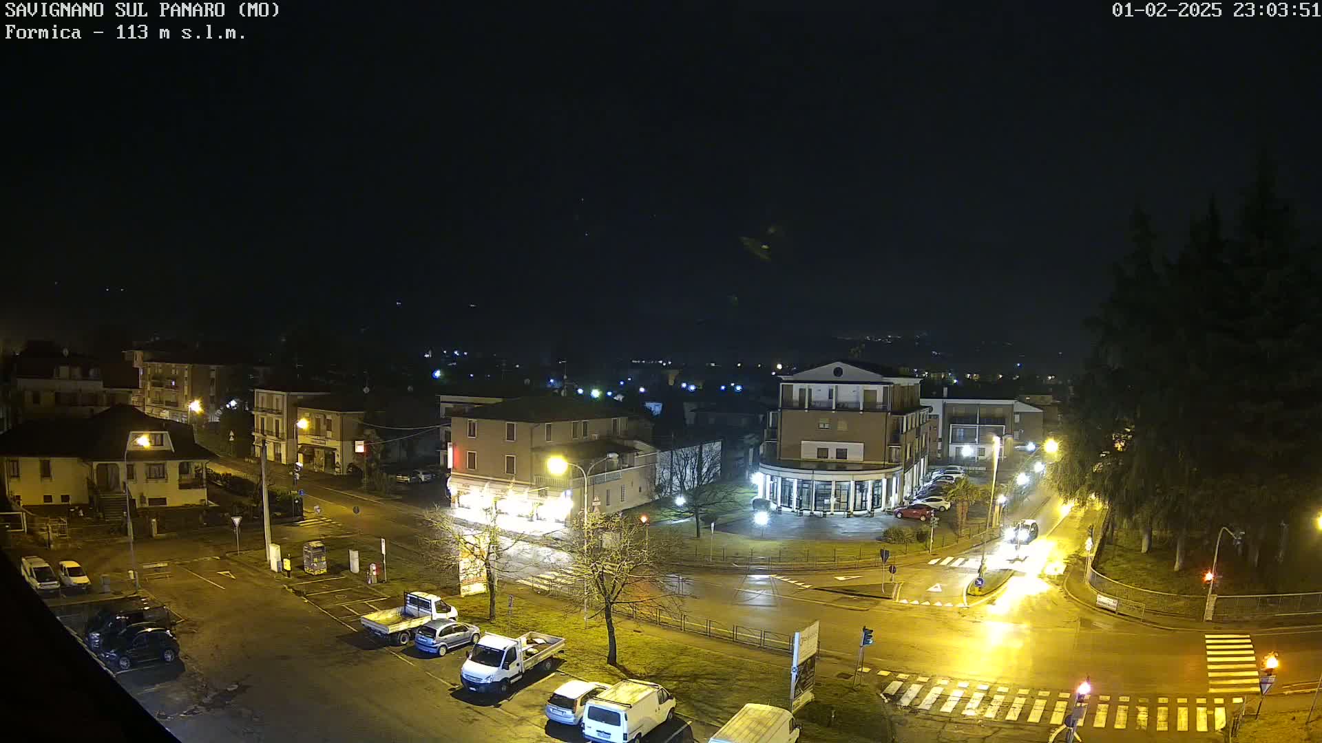 A wet, nighttime street scene shows a town square with parked cars and several buildings illuminated by streetlights.