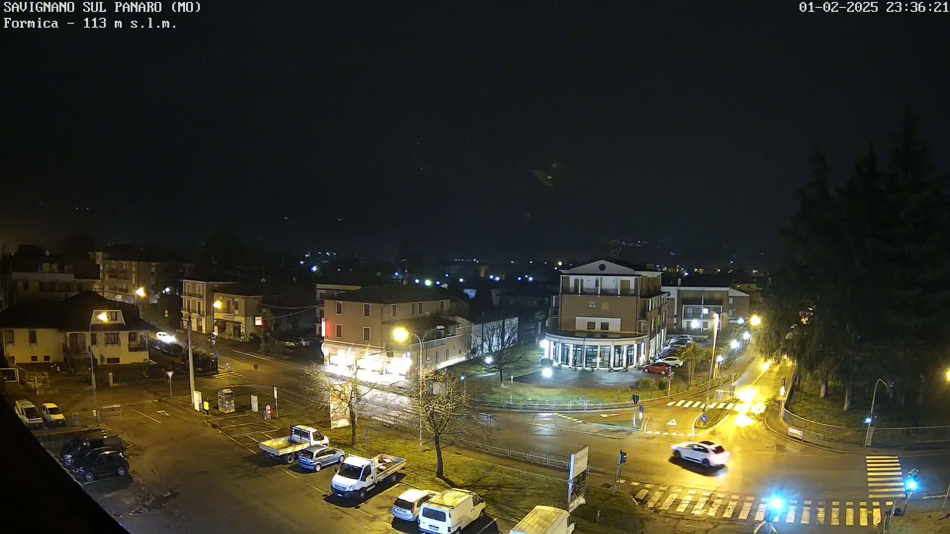 A nighttime, wet view of a town square with parked cars and a few cars driving, surrounded by buildings under a dark sky.