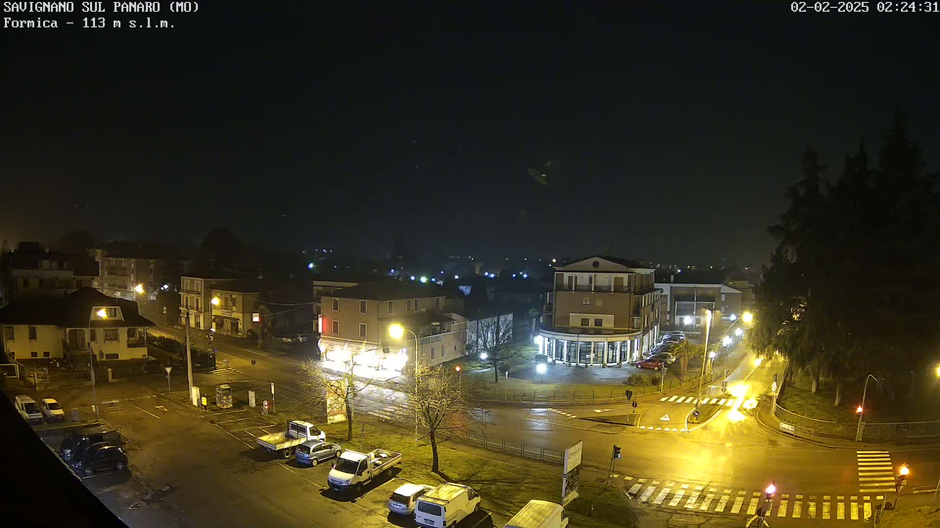 A wet, nighttime view of a town square with parked cars and buildings illuminated by streetlights.