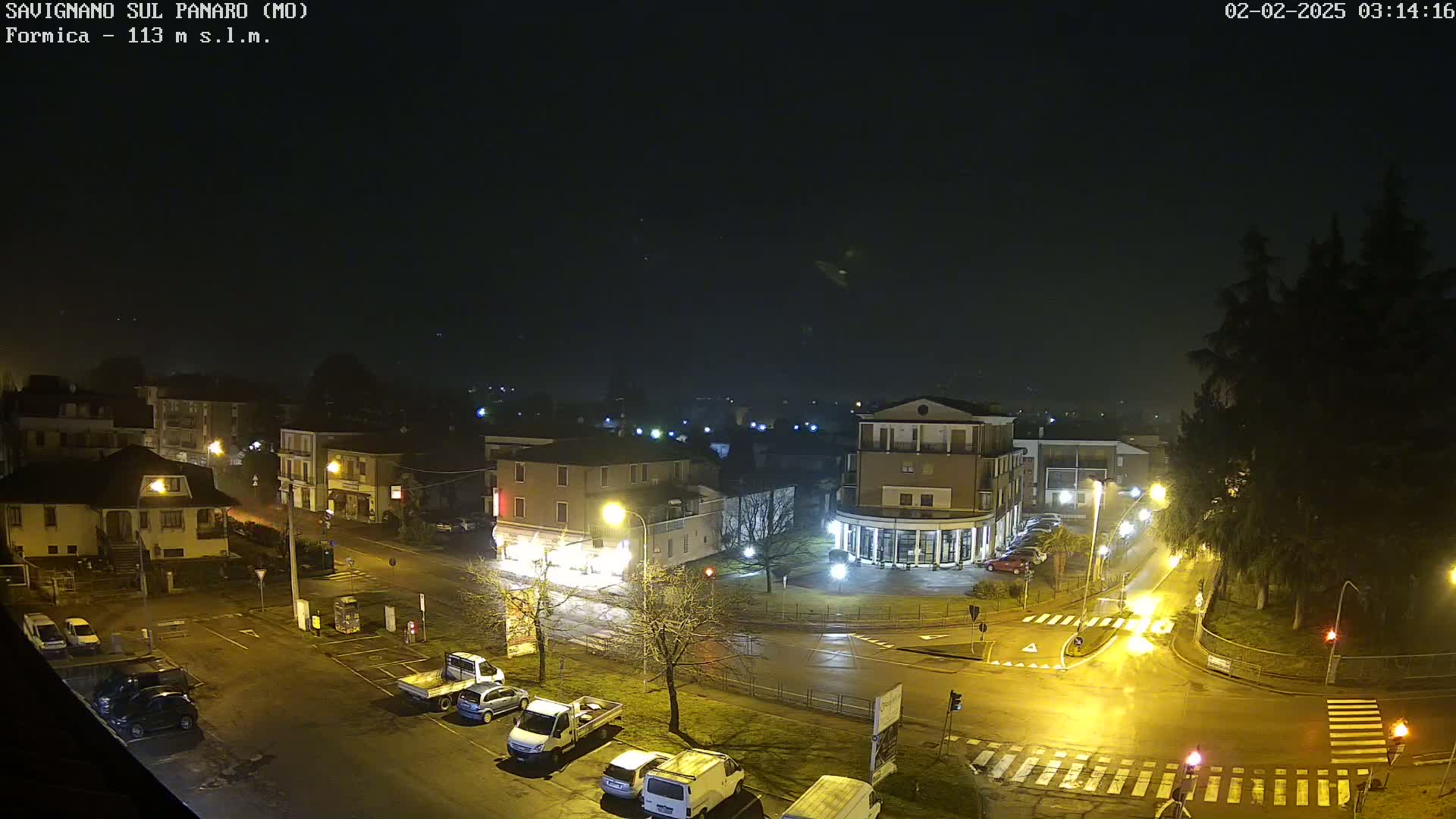 A nighttime, wet scene of a town square with several cars parked and driving, lit by streetlights under a dark, slightly hazy sky.