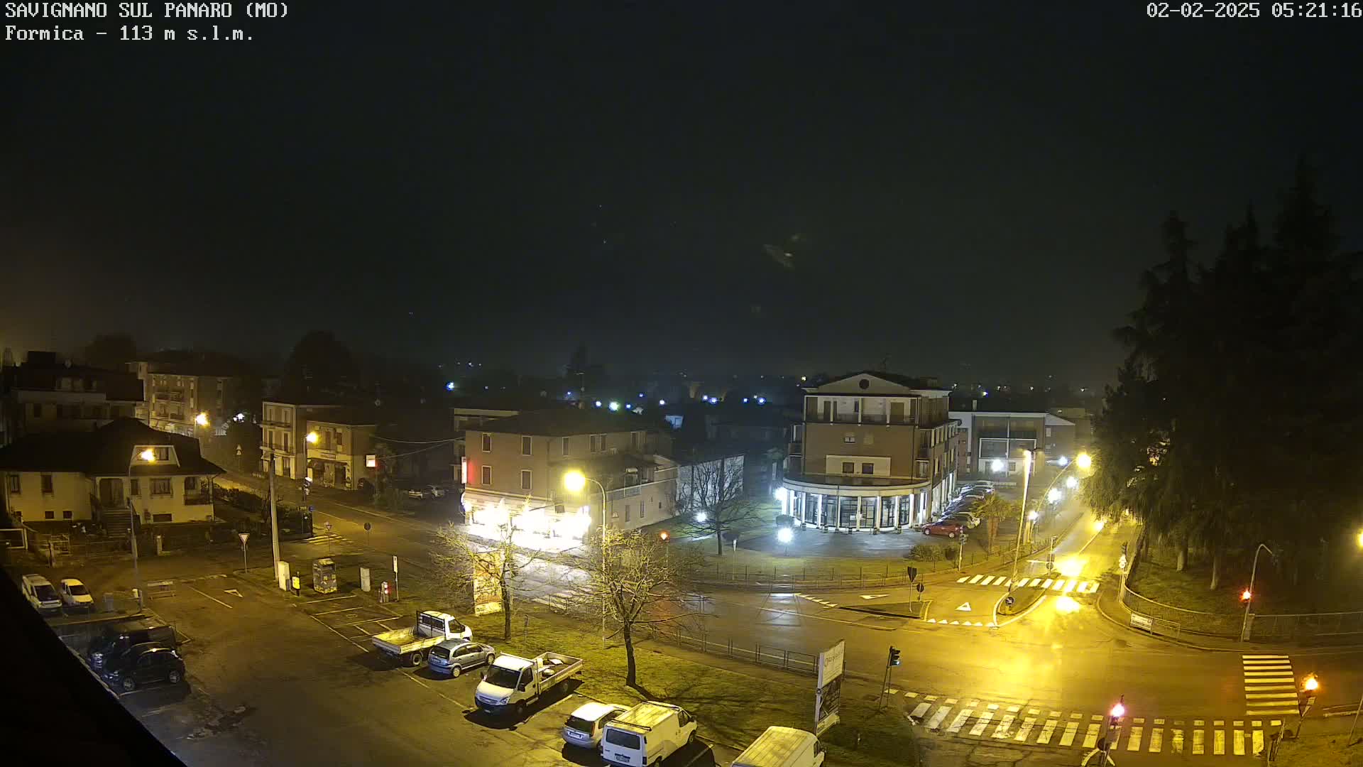 A nighttime, wet view of a town square with several cars parked, streetlights illuminating the area, and buildings visible in the background under a dark, overcast sky.