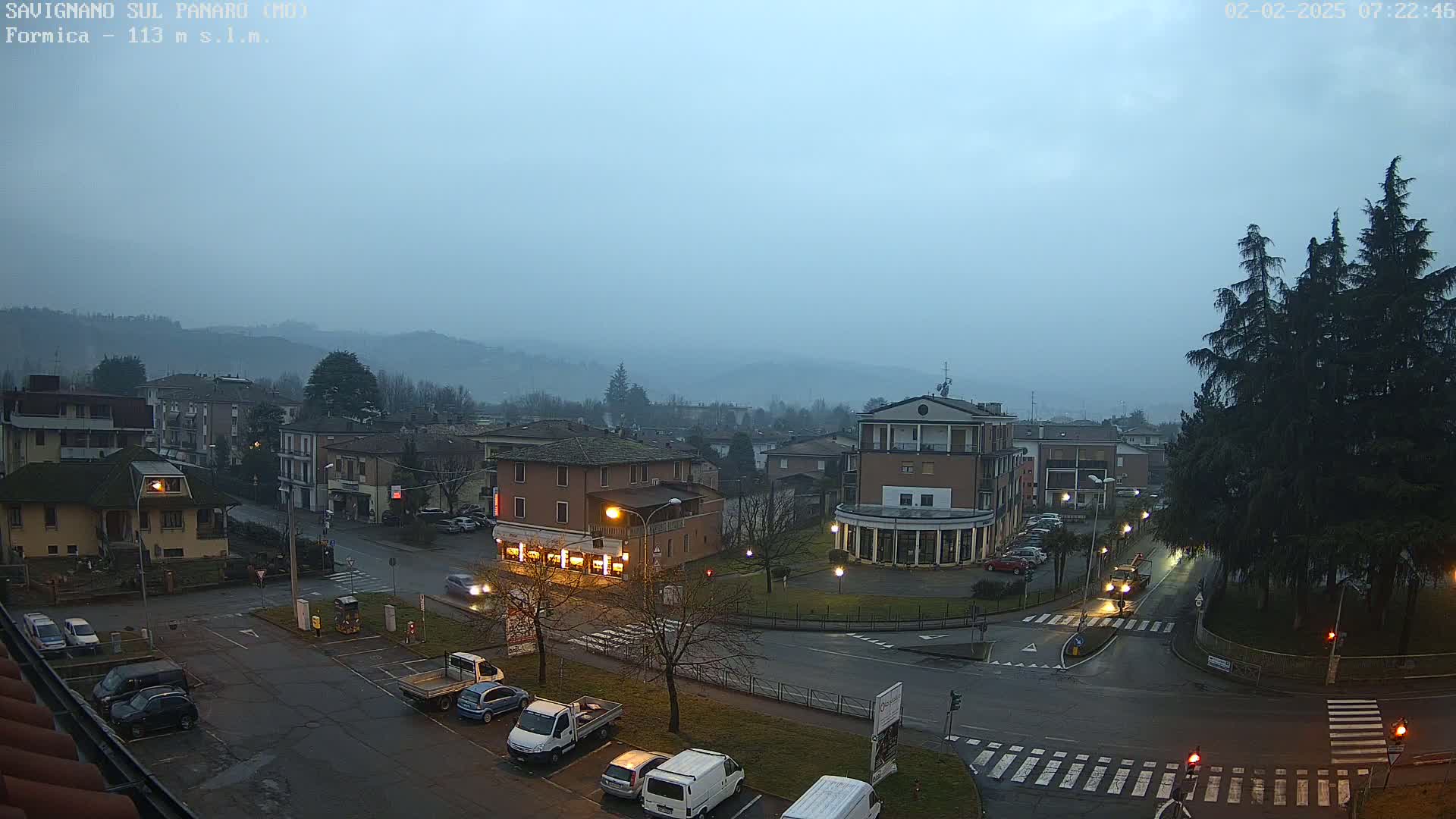 A misty, dimly lit town square with cars parked along the sides of the roads and buildings surrounding the area, under a dusky sky.