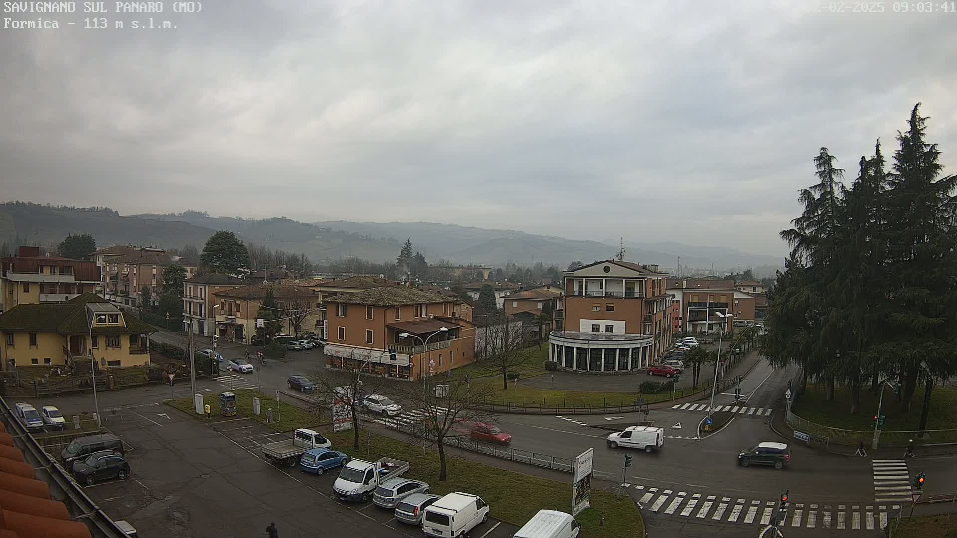 A view of a town square with several buildings, cars, and a pedestrian crossing, set against a backdrop of hazy hills under an overcast sky.