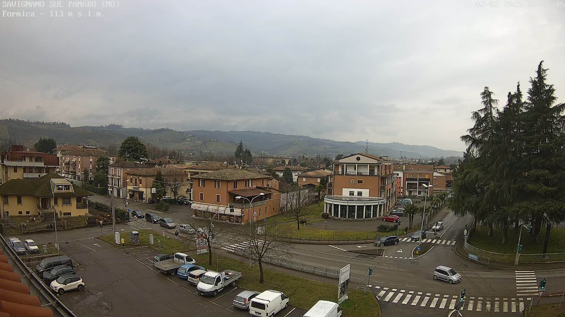 An overcast day reveals a town nestled in a valley, with cars parked in lots and driving on roads that intersect near a small grassy area and larger trees.