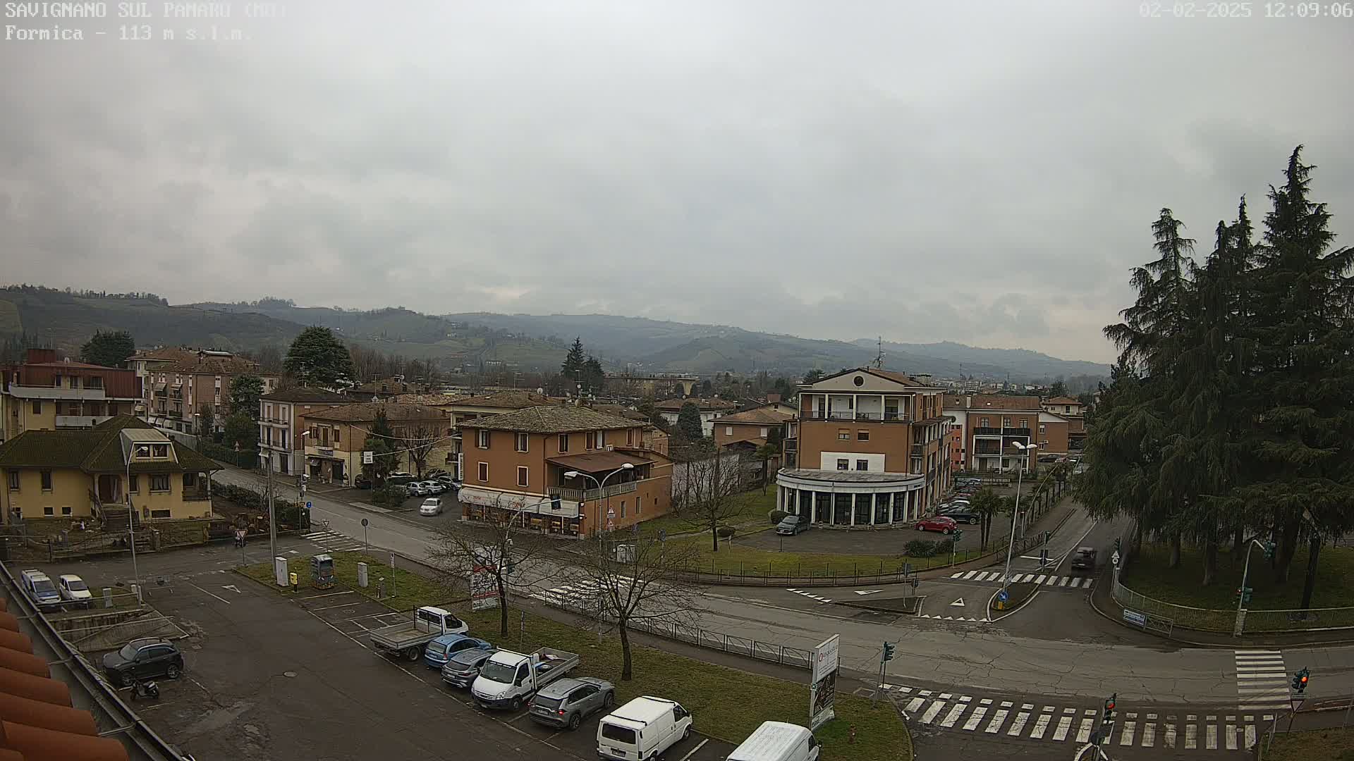 A view of a town situated in a valley under an overcast sky, showing buildings, roads, parked cars, and a few evergreens.