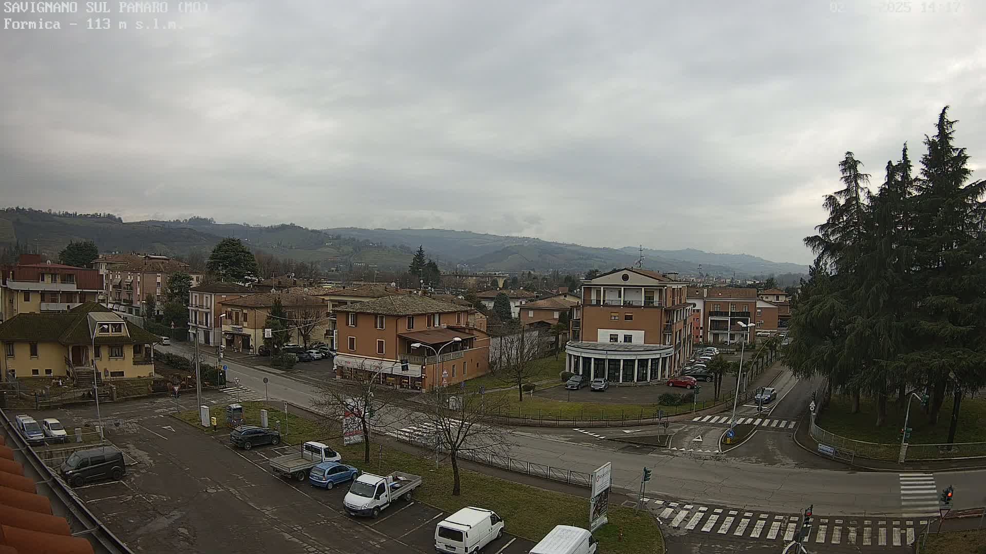 A view of a town nestled in a valley under a cloudy sky, with several buildings, cars parked in lots, and evergreen trees.