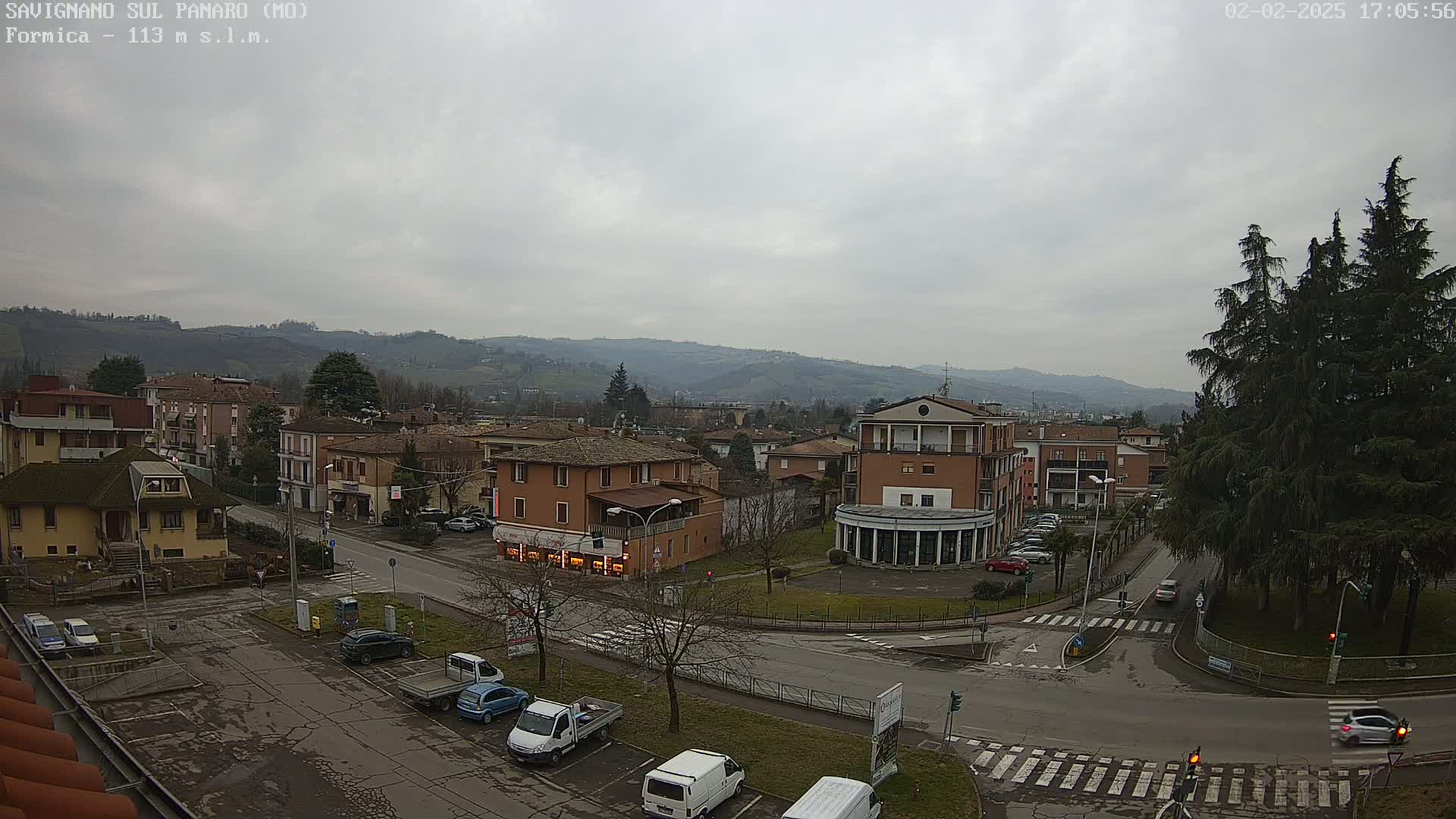 A view of a town nestled in a valley under an overcast sky, showing buildings, roads, parked cars, and evergreen trees.