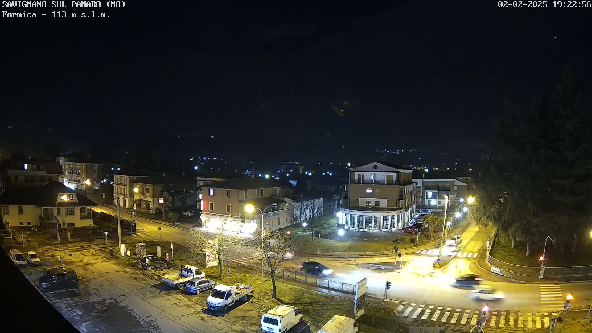 A nighttime aerial view of a town with a few cars on the roads, slightly wet roads, and streetlights illuminating the scene under a clear, dark sky.