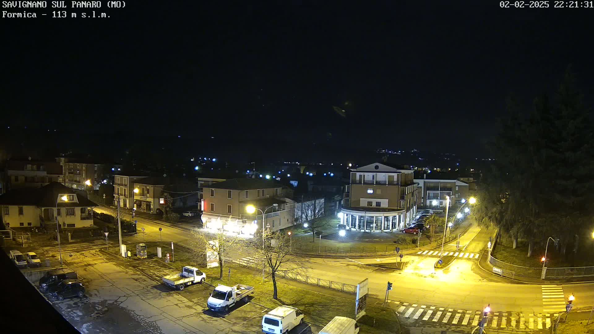 A nighttime, high-angle view of a town square with parked vehicles and illuminated buildings under a clear, dark sky.